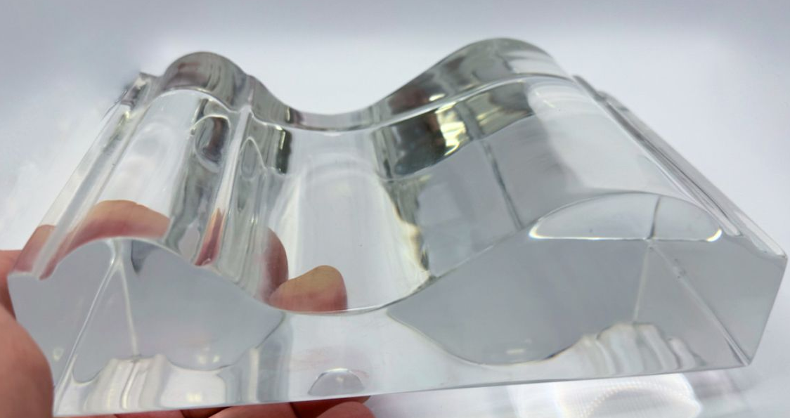 A hand holds a thick, clear acrylic piece with a wavy, molded top surface against a plain white background.