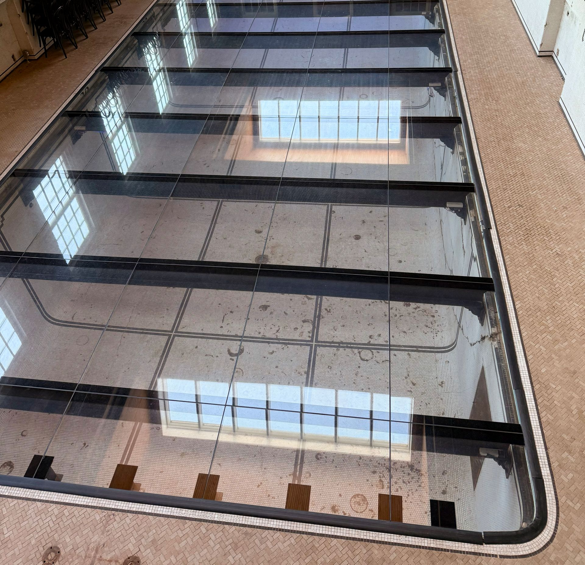 Glass floor with rectangular panels, set in a histpric pool in a light brown room. Black supports frame the glass panels.