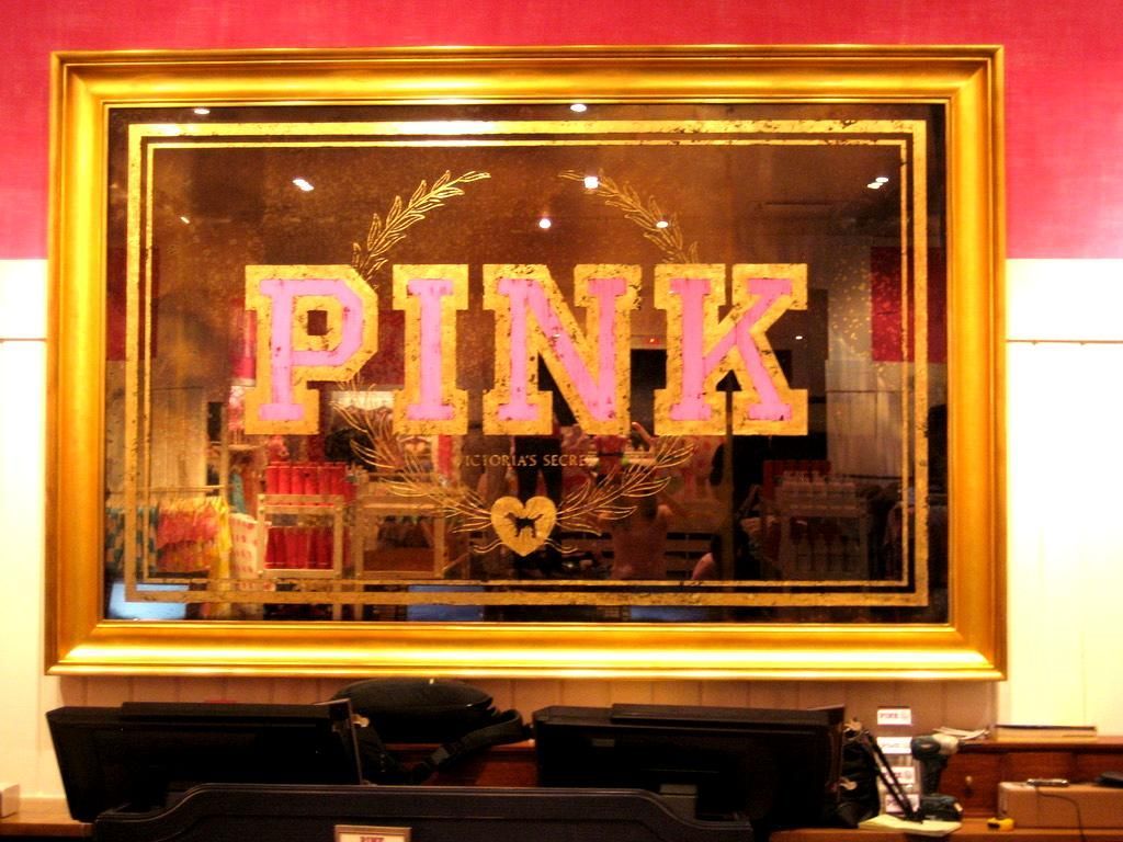 Pink Victoria's Secret store mirror sign, gold frame, pink logo, store interior with checkout counter visible.