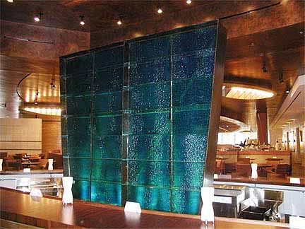 A large teal-colored textured glass feature in a restaurant. It is made of square panels and set behind a bar.
