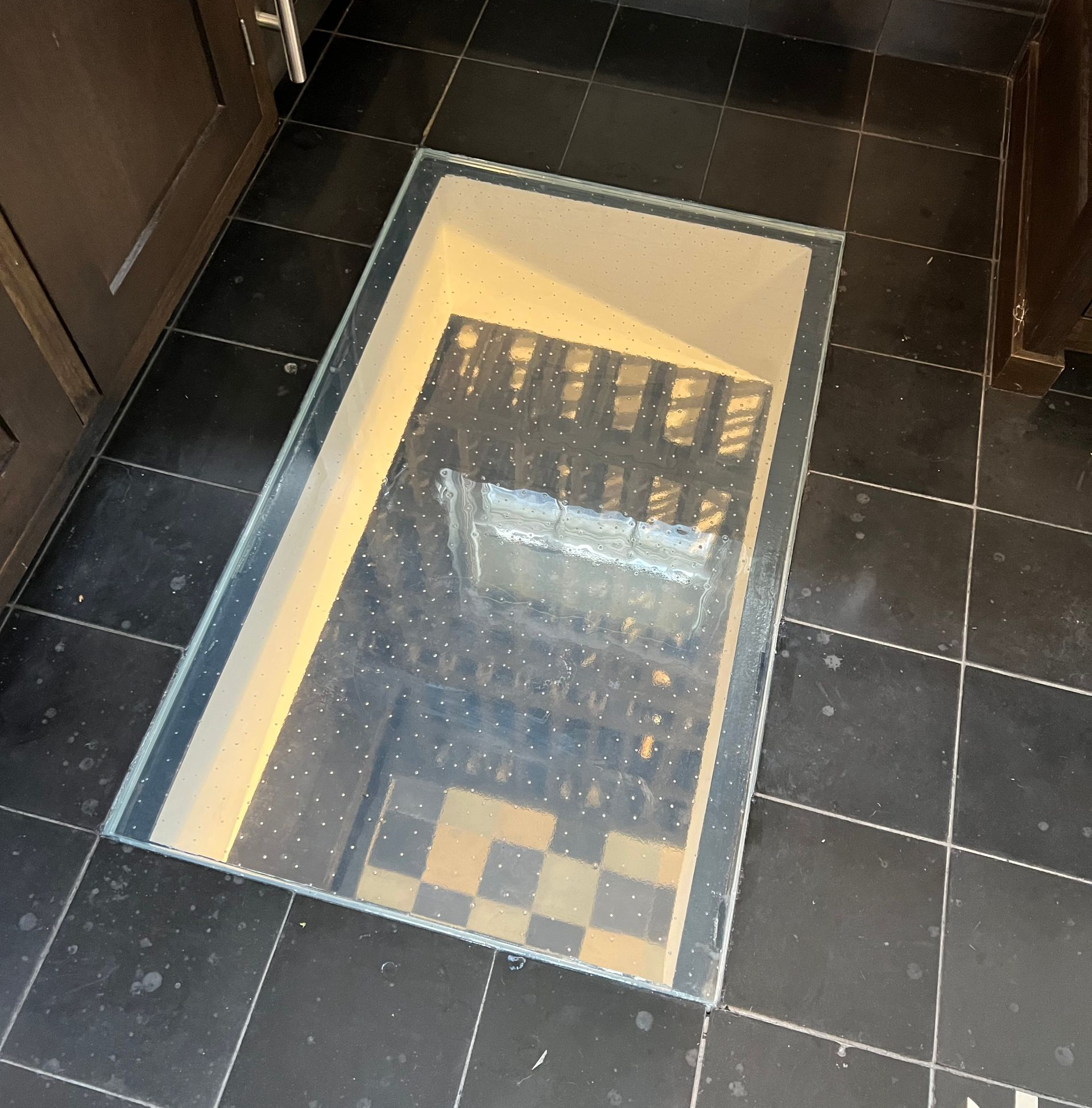 Glass floor covered cellar access in black-tiled floor.