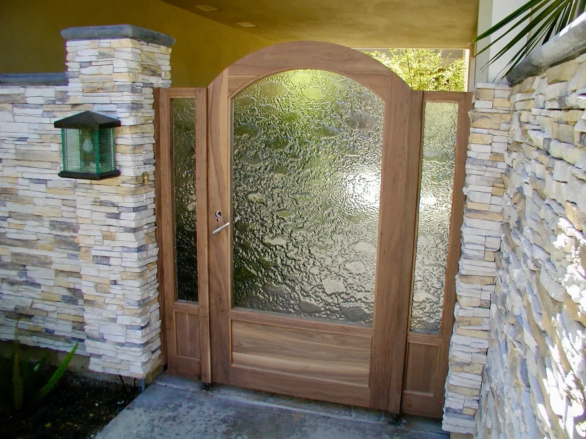An arched wooden gate with textured glass panels set between two stone pillars and walls.