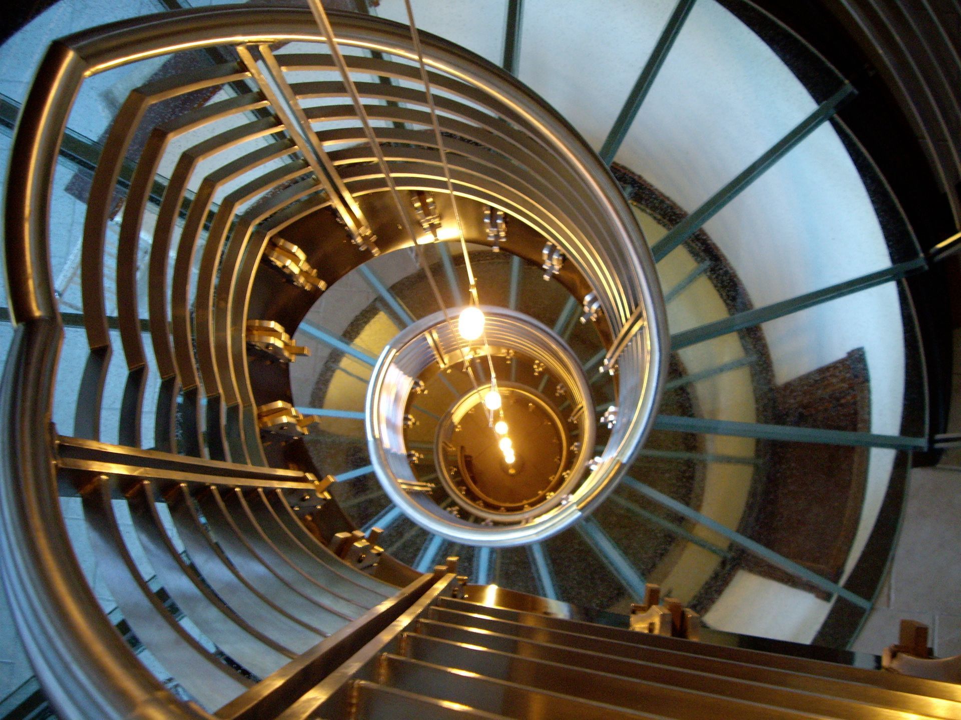 Spiral staircase, stainless steel railing, leading upwards, with overhead lighting.