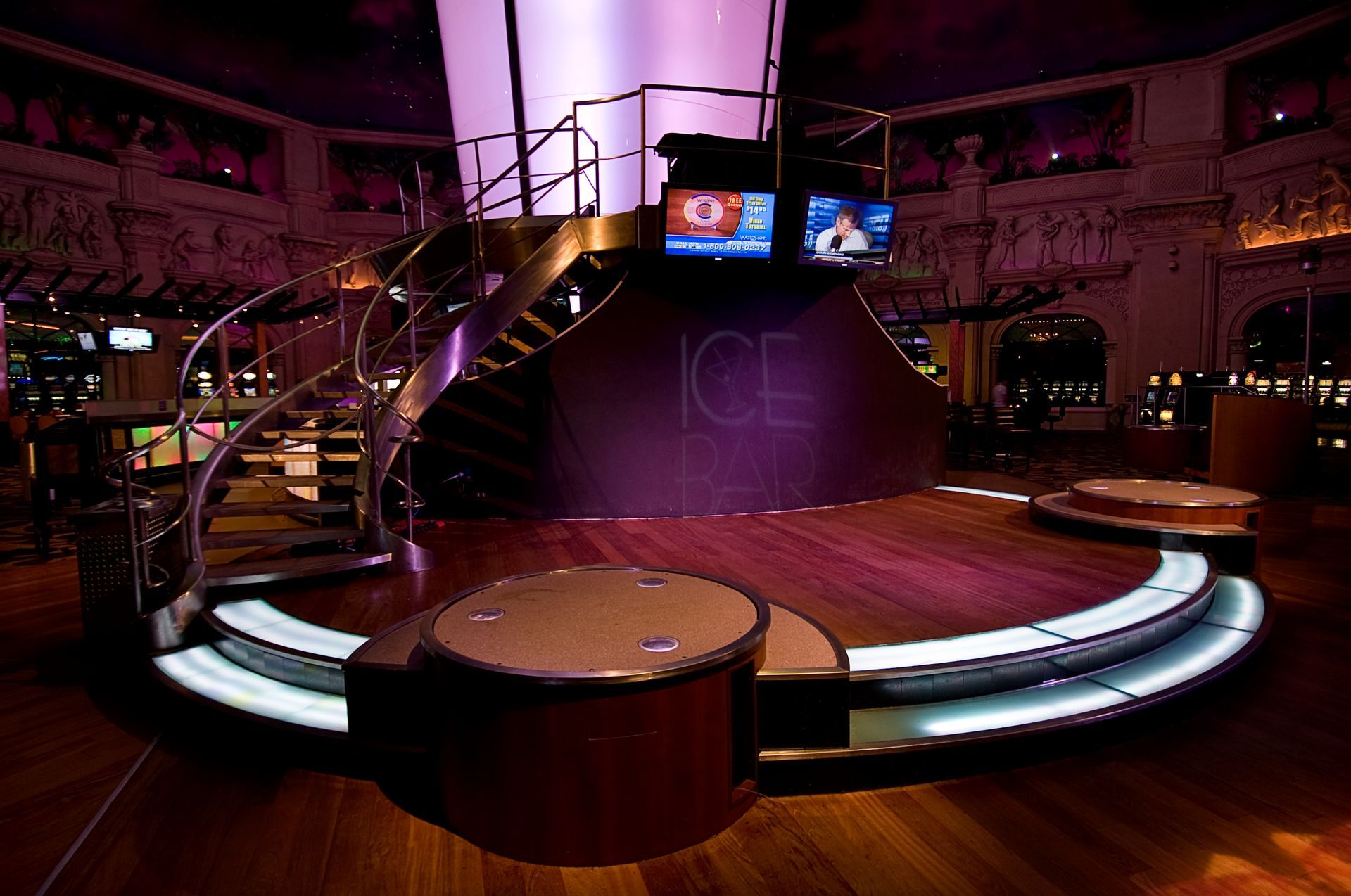 A dark, elegant lounge features a circular central stage with steps, lit by purple ambient light and two small screens.