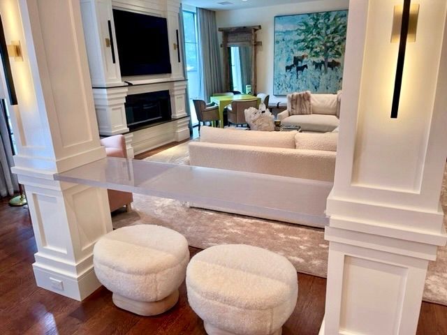 Living room with white columns, sofa, fireplace, and painting. Two round cream stools in front of a clear desk.