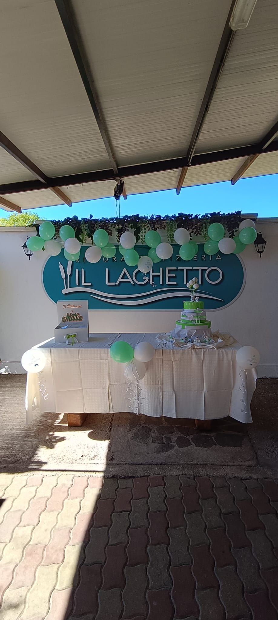 Un tavolo addobbato per una festa con una tovaglia bianca, una torta a più piani e un arco di palloncini sullo sfondo di un'insegna con la scritta