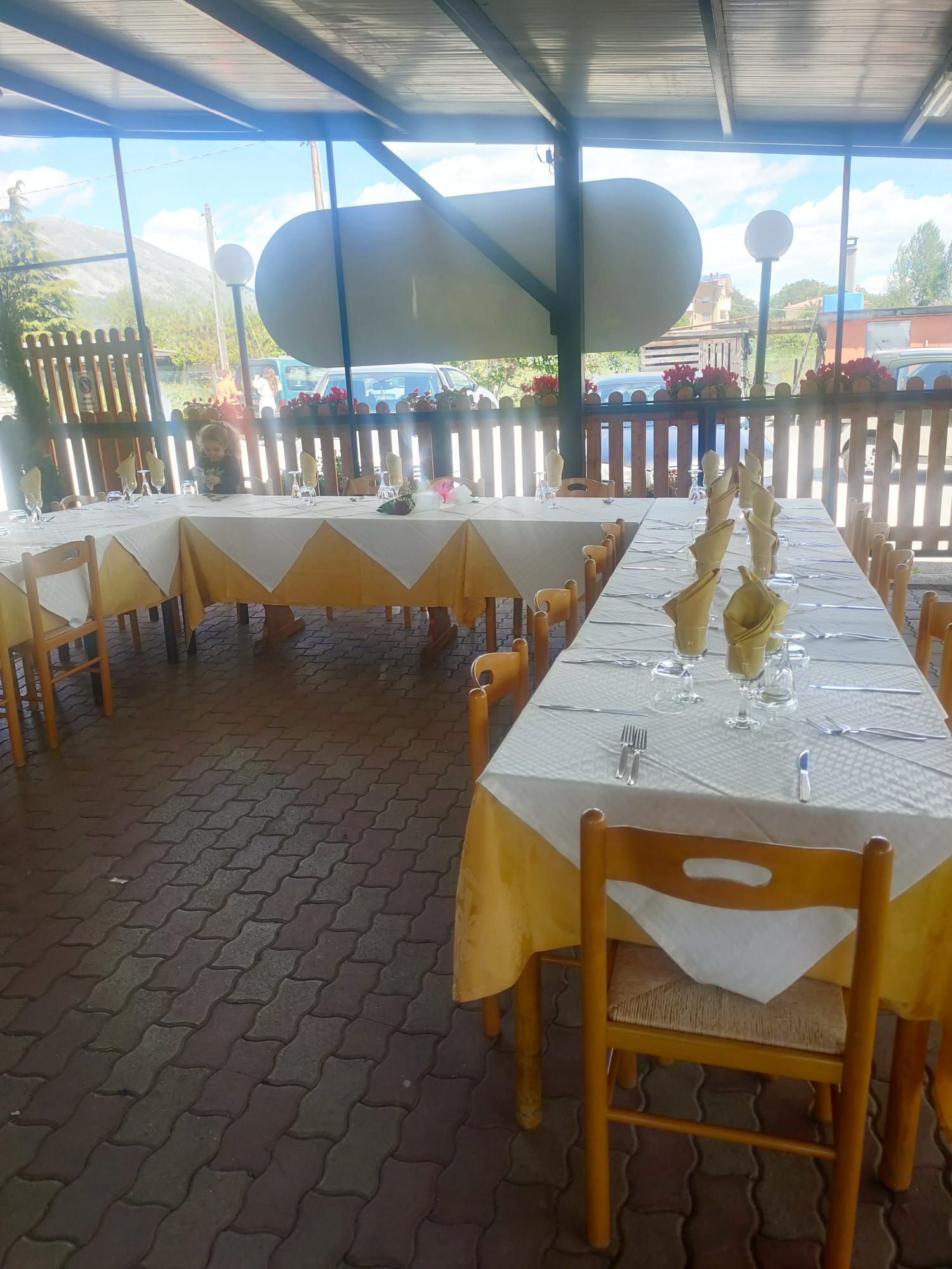 Tavoli apparecchiati con tovaglie bianche e gialle, pronti per un pasto sotto un patio esterno coperto da una recinzione di legno.