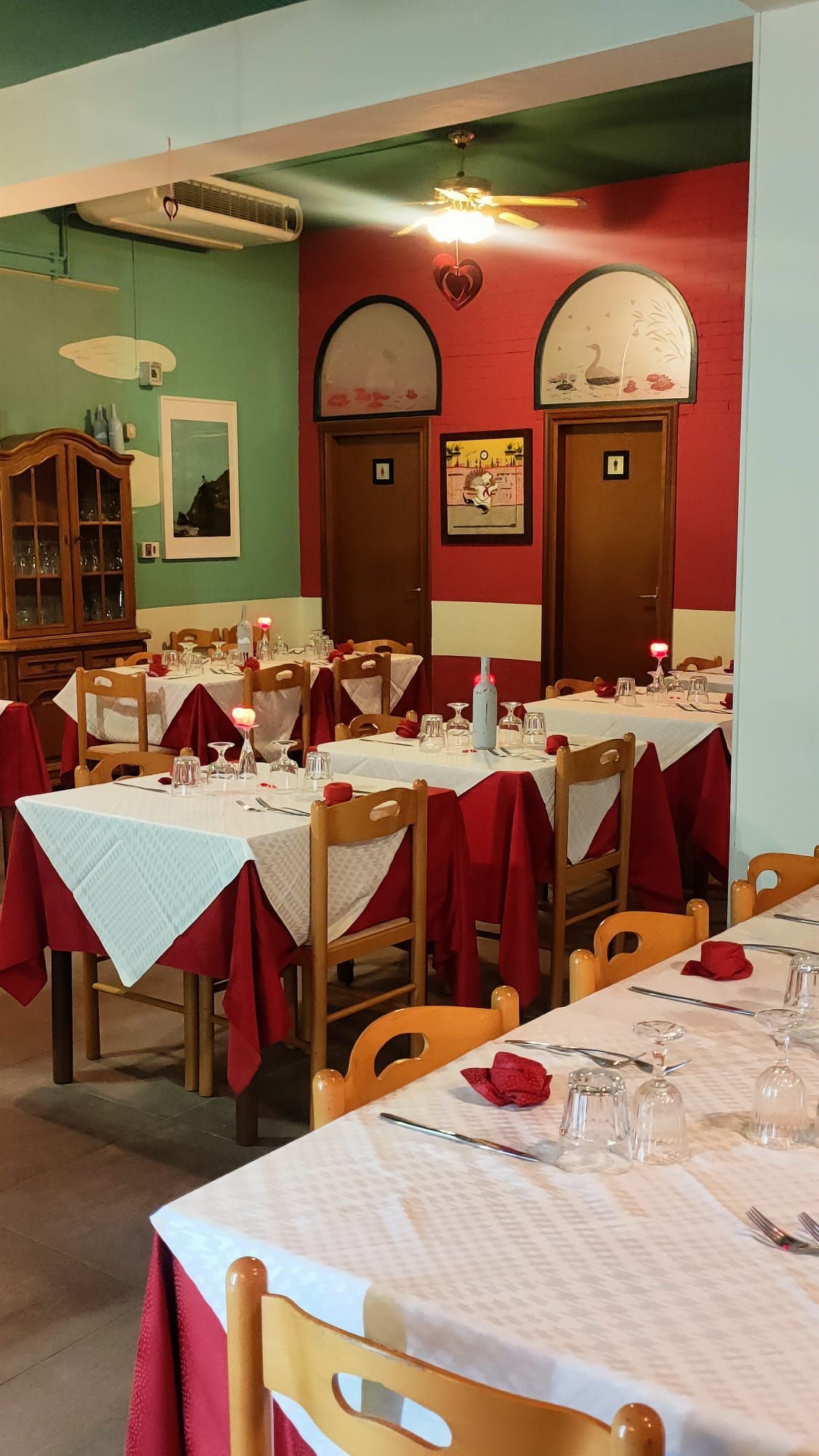 Interno di un ristorante con tavoli apparecchiati per la cena, con tovaglie bianche, copritovaglie rosse e sedie in legno.