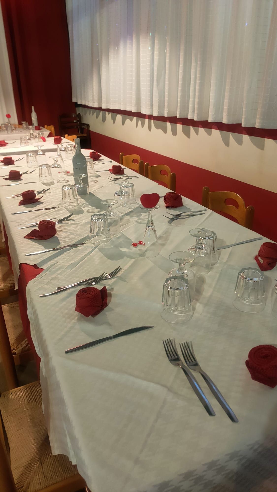 Un lungo tavolo da pranzo apparecchiato per una cena formale con tovaglie bianche, posate, bicchieri d'acqua e rose rosse decorative.