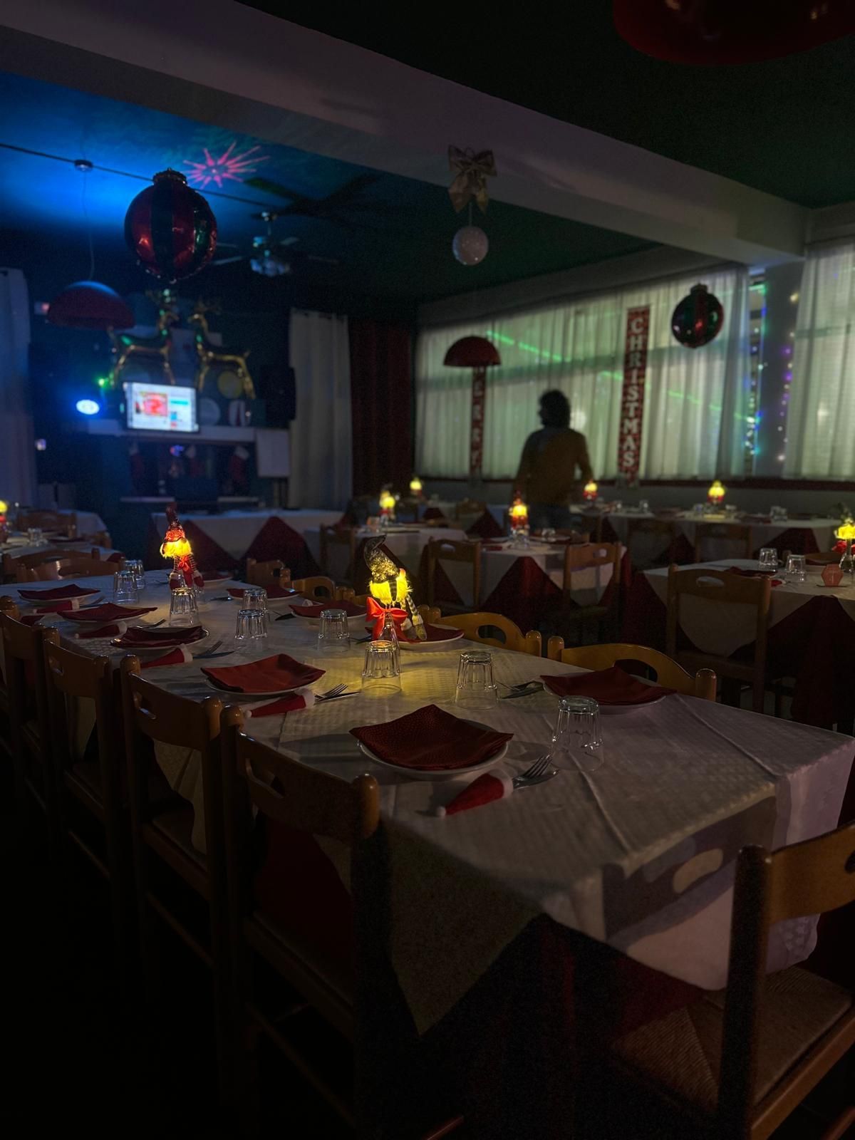 Una sala da pranzo in penombra, apparecchiata per un'occasione festiva con lunghi tavoli, tovaglie bianche, tovagliette rosse e luci calde.