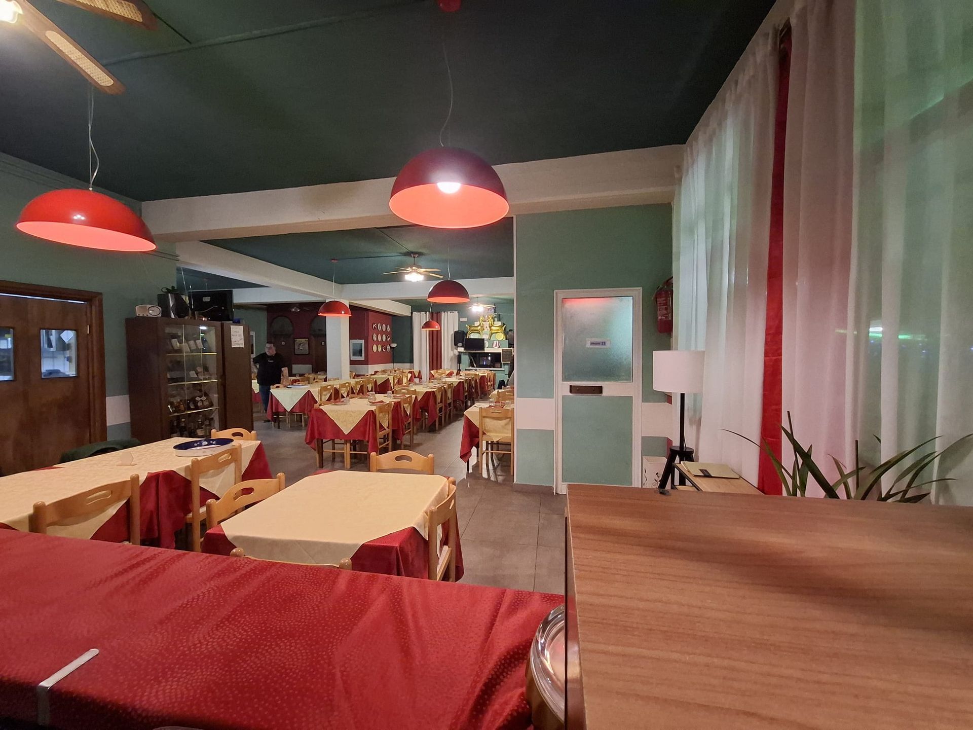 Una sala da pranzo con tavoli ricoperti da tovaglie beige e copritovaglie rosse, illuminata da lampade a sospensione rosse sotto un soffitto scuro.