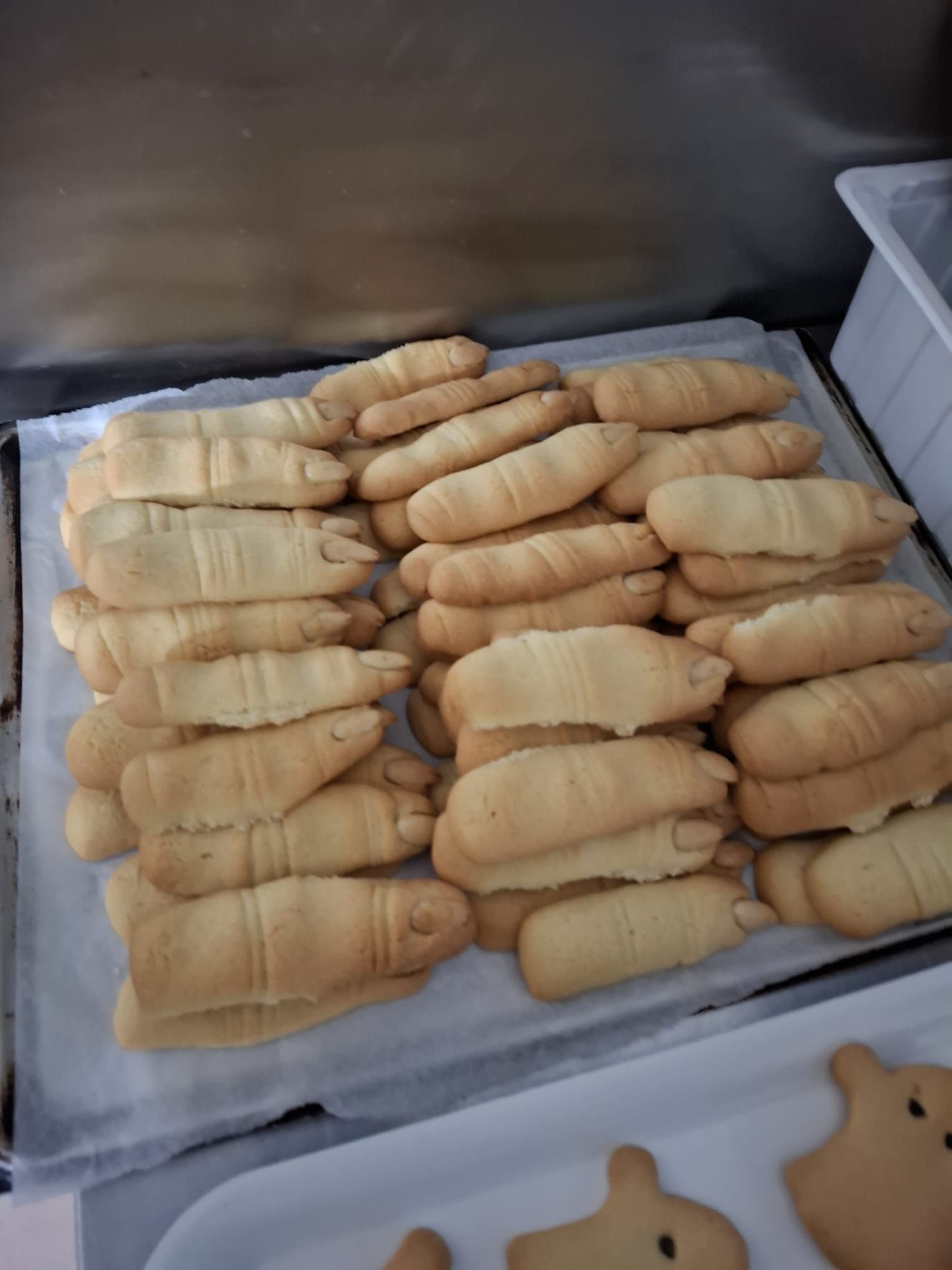 Una teglia da forno piena di biscotti chiari a forma di dito, disposti in file, con biscotti a forma di fantasma visibili sotto.