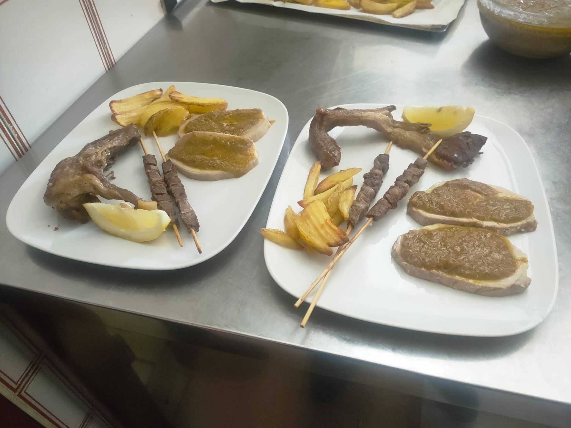 Due piatti bianchi su una superficie in acciaio inossidabile, ciascuno contenente spiedini, spicchi di patate, fette di pane spalmato e uno spicchio di limone.