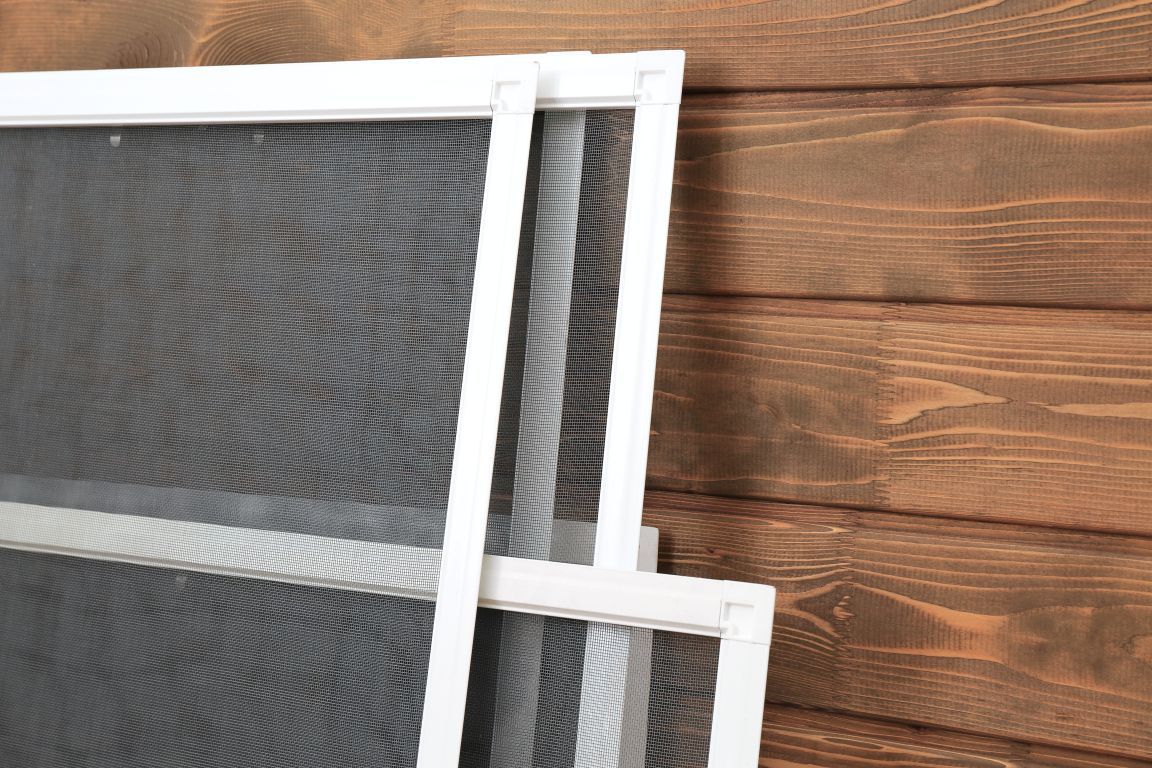 Stack of white-framed window screens with gray mesh, leaning against a brown wooden wall.