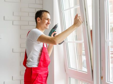 A man in red overalls is installing a window with a drill.