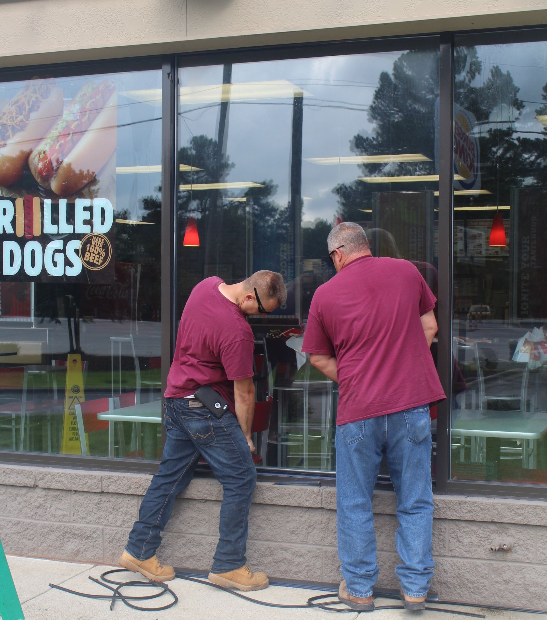 Two men in maroon shirts installing a window at a restaurant with grilled dogs advertisement.