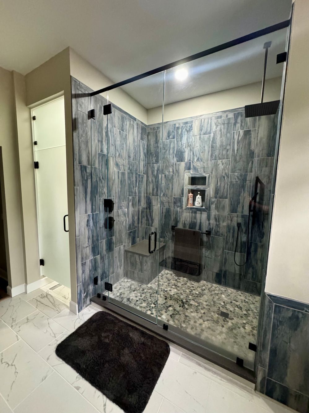 Modern shower with blue-grey tiled walls, a glass door, and a pebble floor. A black bath mat is on the floor.