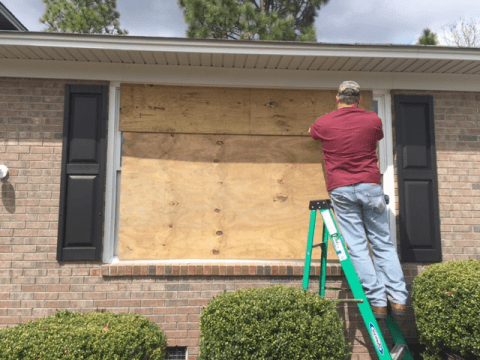A man is standing on a ladder covering a window with plywood.
