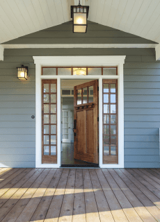 Wooden door open on a gray house porch with side windows and lamps.