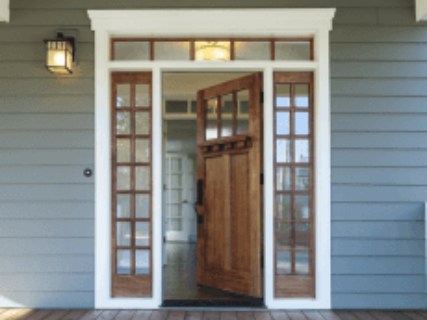 Wooden front door open, flanked by glass panel windows, set in a gray-blue house with white trim.