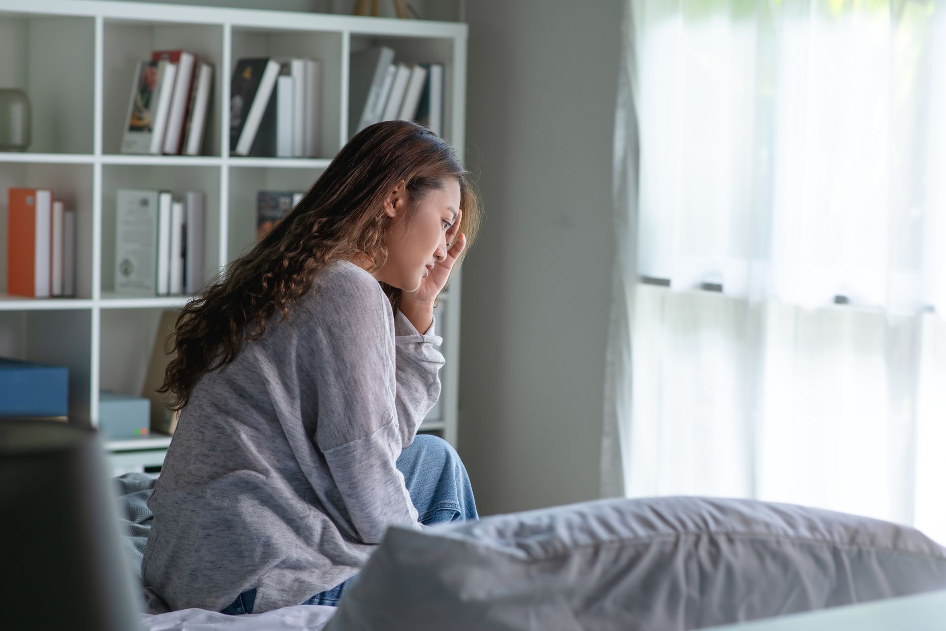 Woman sitting on a bed, looking distressed. She holds her head near a window and bookshelf. Woman sitting on a bed, looking distressed. She holds her head near a window and bookshelf.