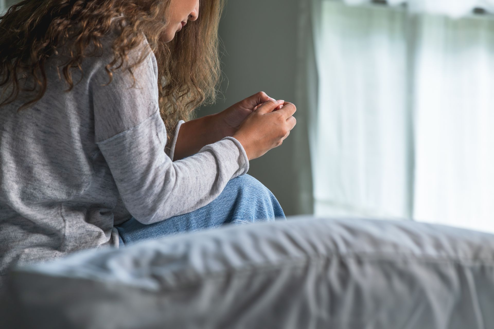 Woman sitting on bed, head down, hands clasped. Natural light from window. Woman sitting on bed, head down, hands clasped. Natural light from window.