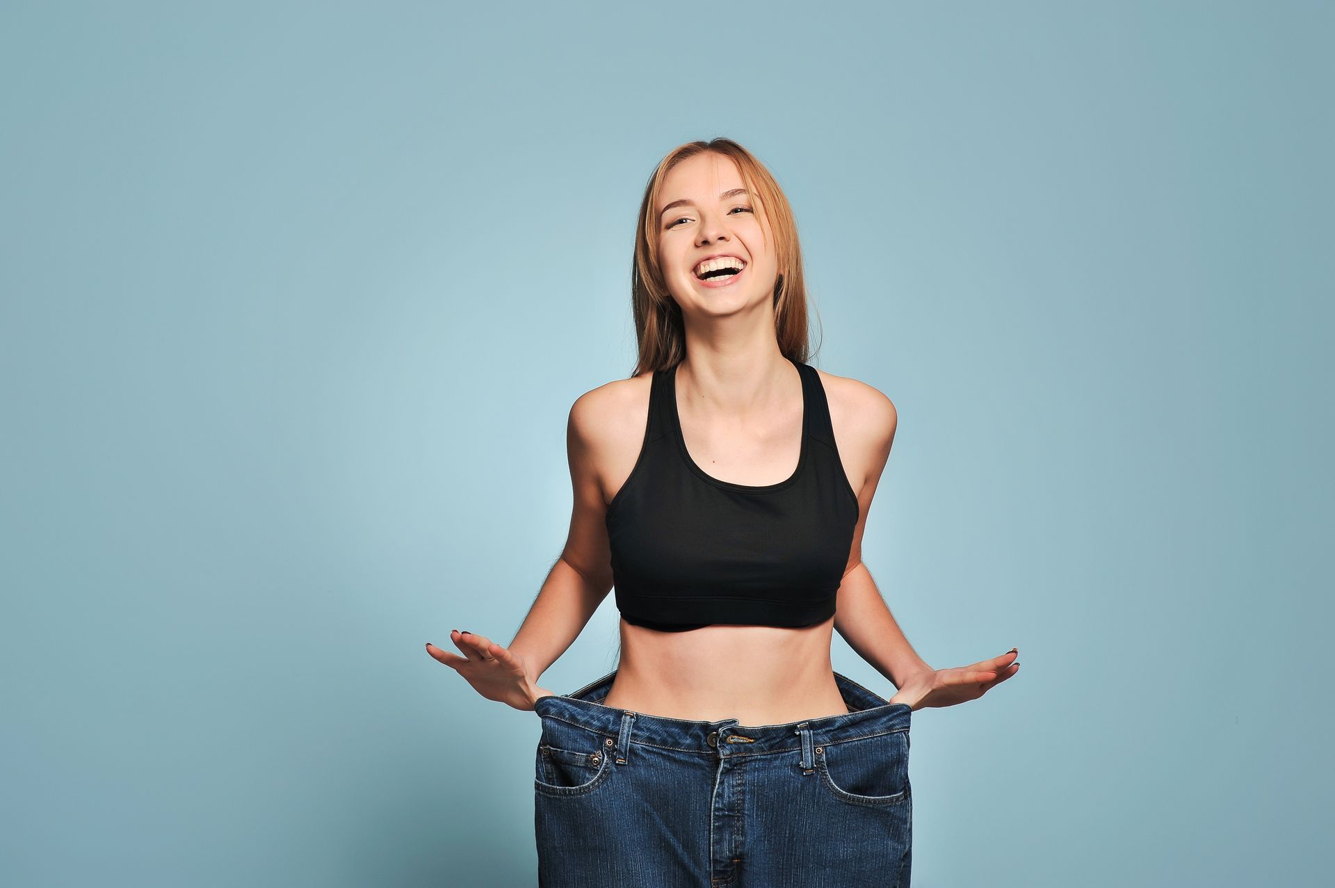 Woman wearing oversized jeans, smiling, holding out sides, showing weight loss. Light blue background.