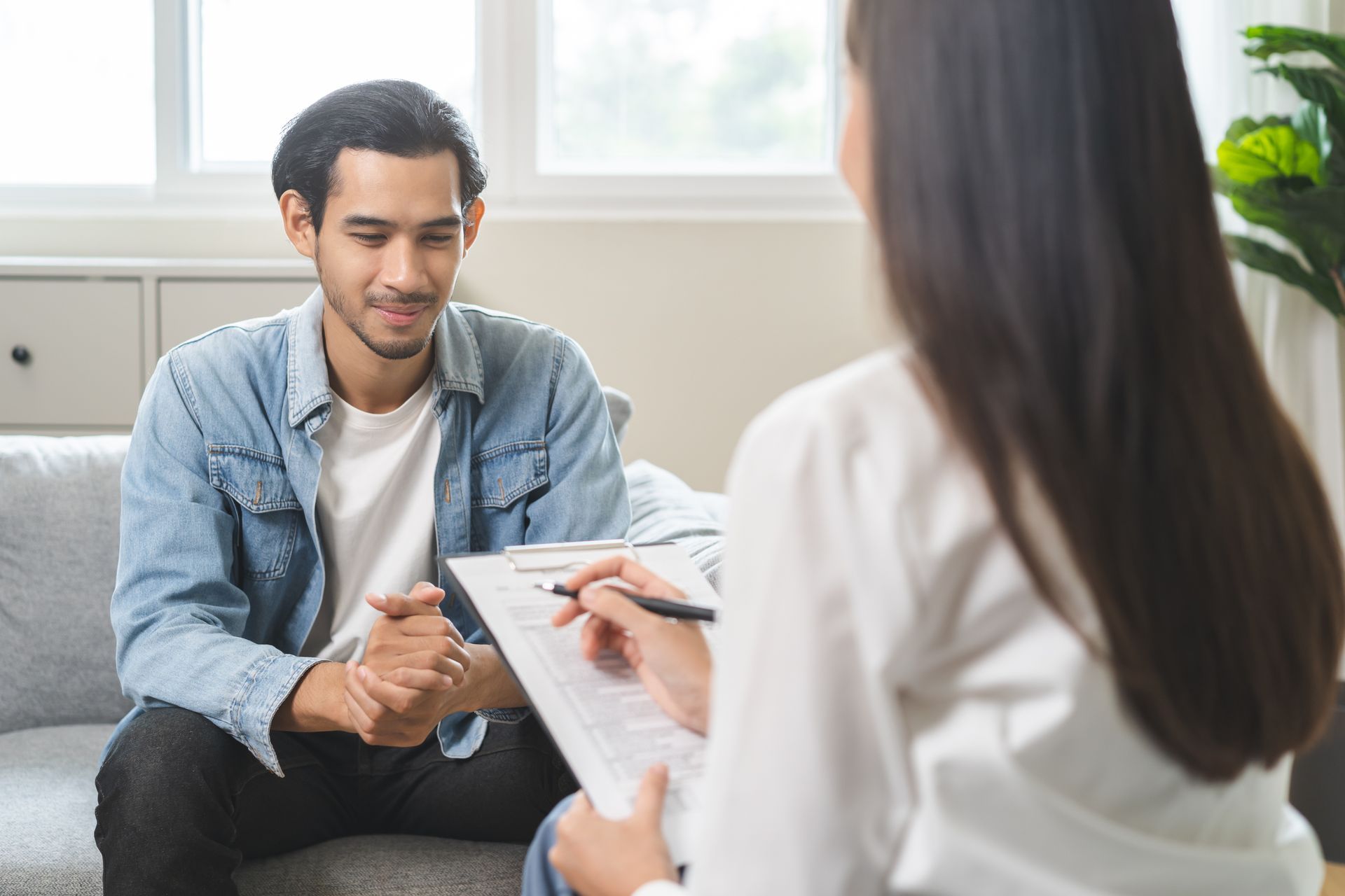 Man sits on a sofa, looking at a person with long hair writing on a clipboard in a room. Man sits on a sofa, looking at a person with long hair writing on a clipboard in a room.