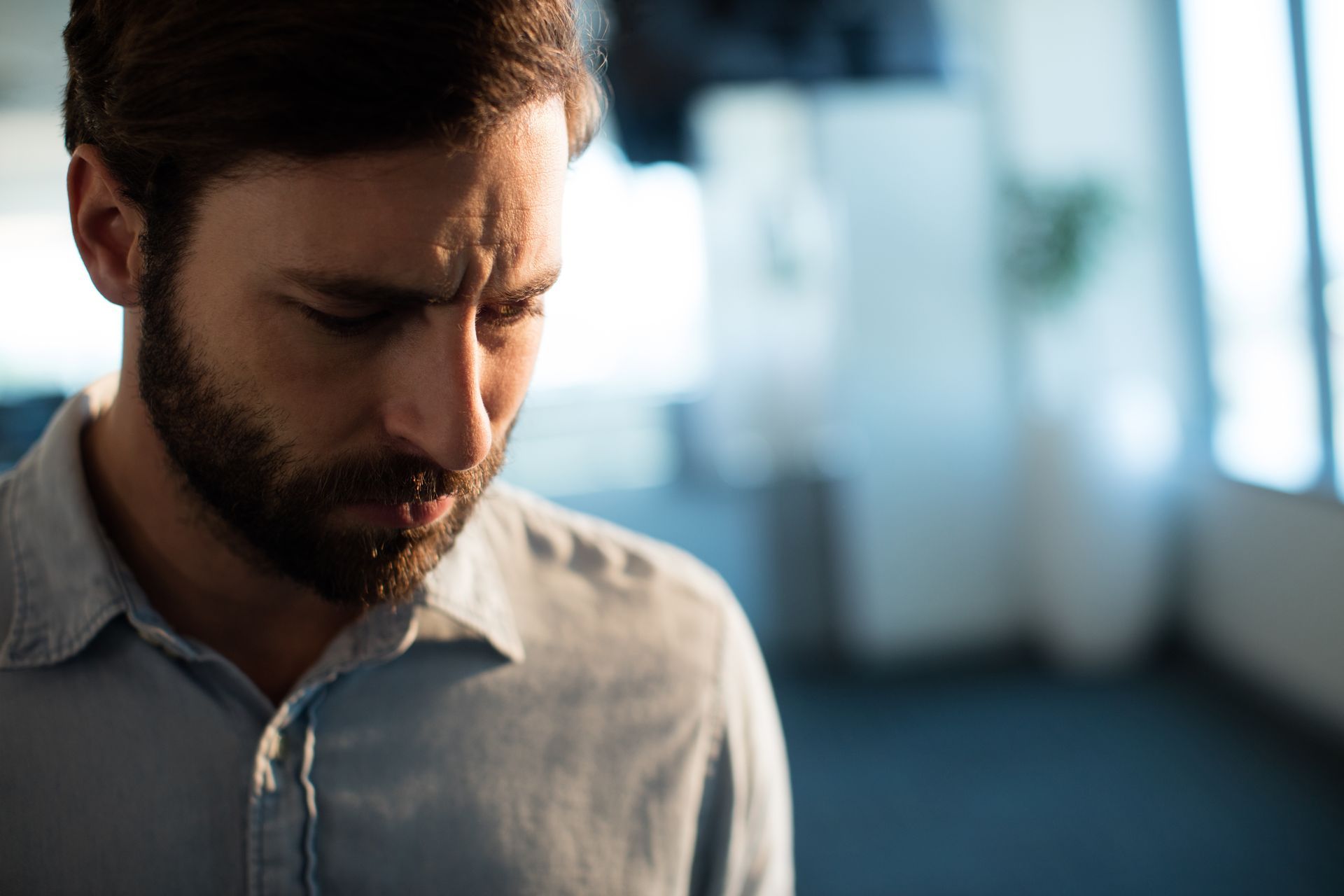 Man with beard in light blue shirt, looking down with a concerned expression. Man with beard in light blue shirt, looking down with a concerned expression.