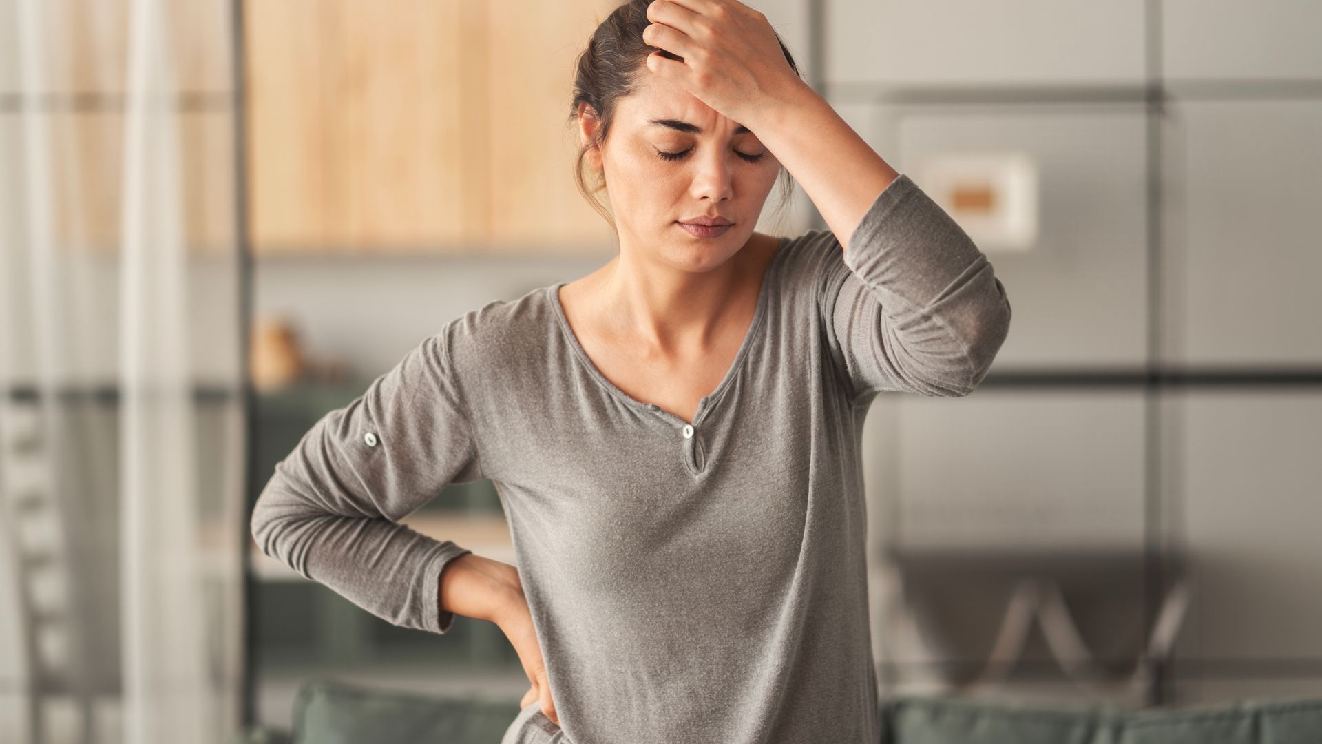 Woman holding her head and back, appearing to be in pain, indoors.