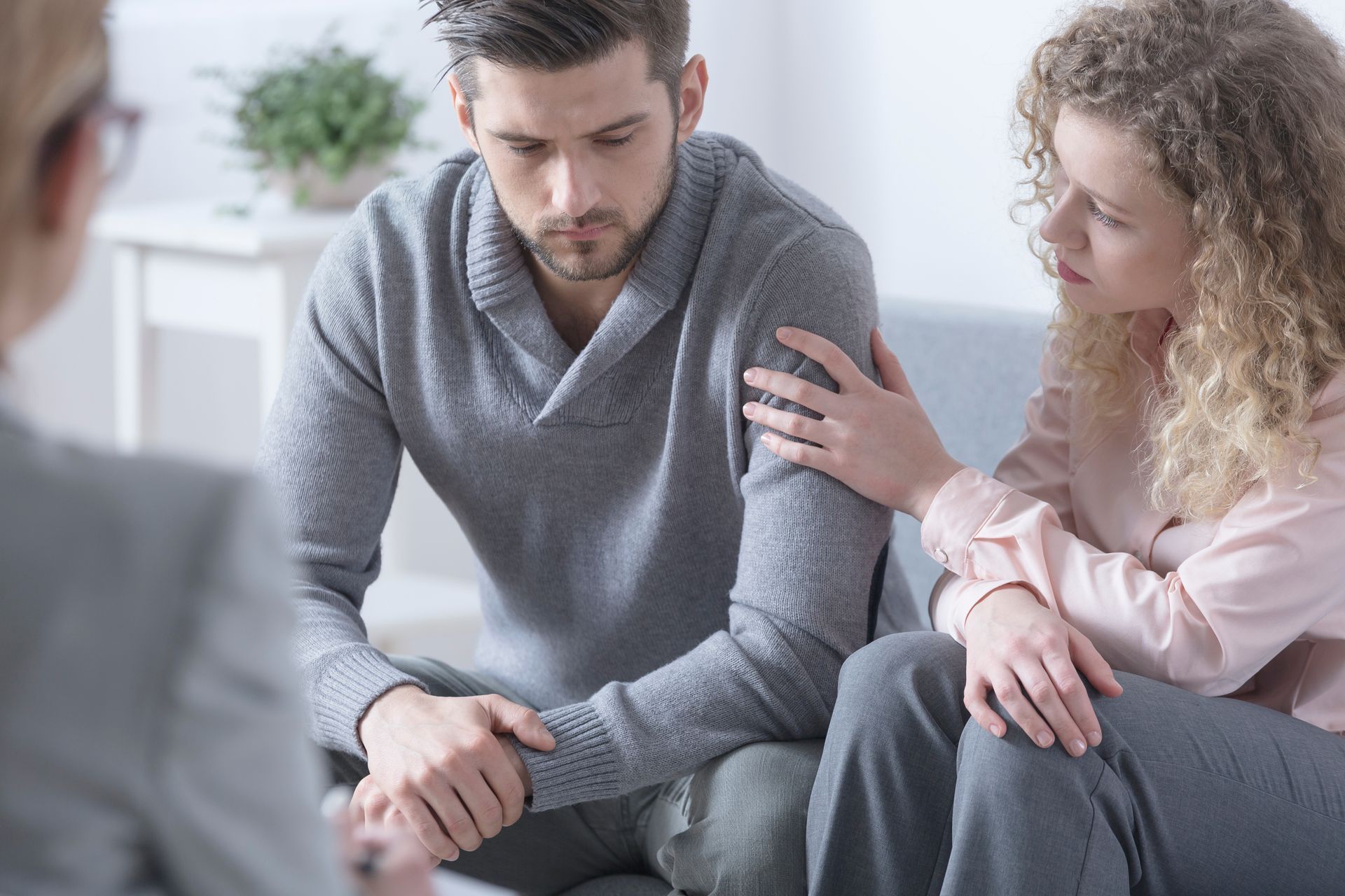 Couple in therapy session, woman comforts man, seated, therapist observes.