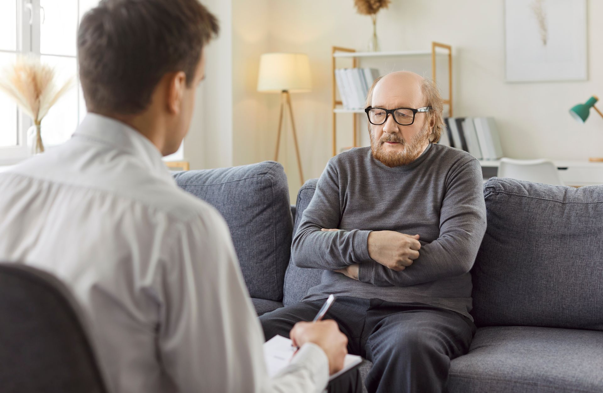 Therapist taking notes during session with a patient on a couch. The patient has glasses and arms crossed. Therapist taking notes during session with a patient on a couch. The patient has glasses and arms crossed.