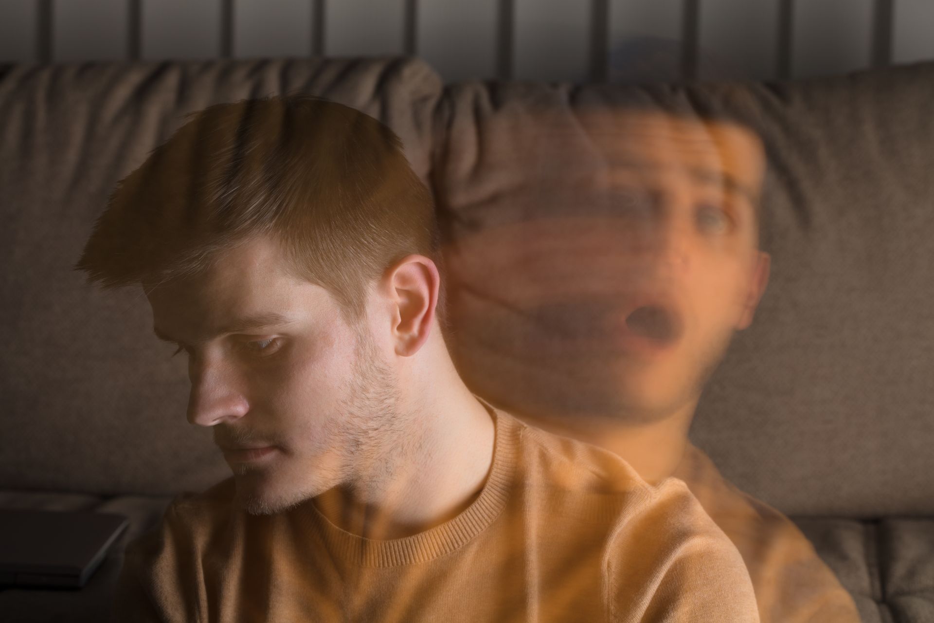 Man with blurred double exposure effect, looking down with a concerned expression, sitting on a couch.