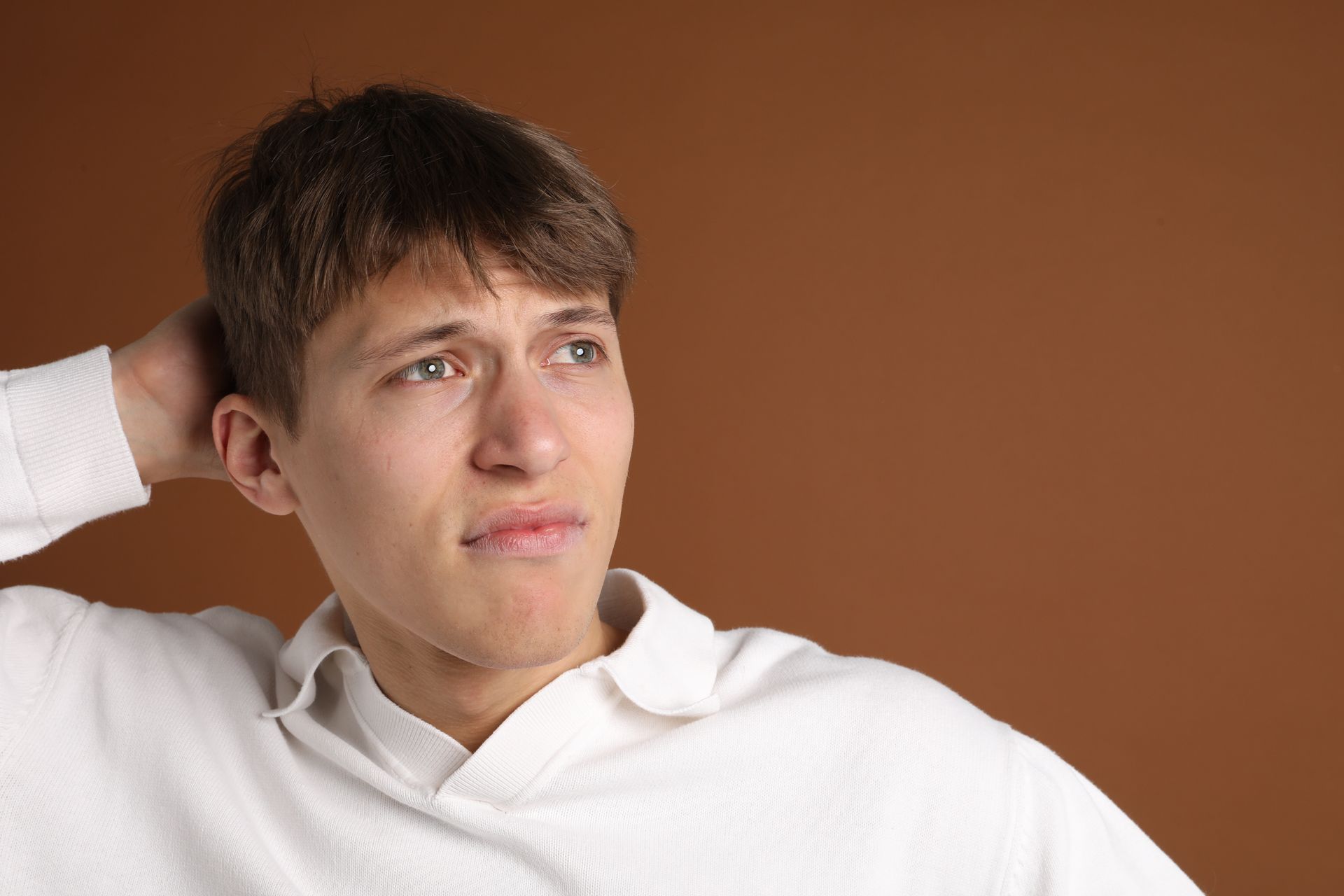 Man in white sweater, hand behind head, looking off to the side with a contemplative expression against a brown background.