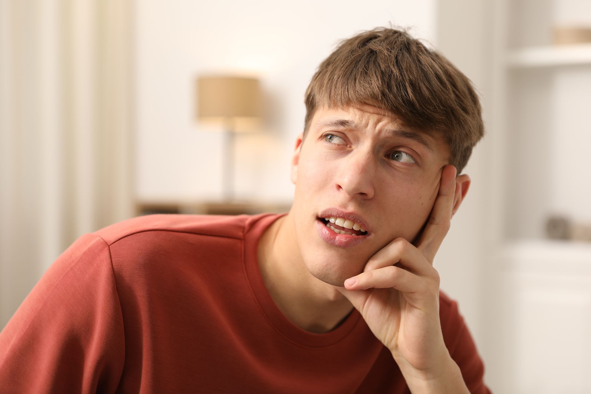 Man in a red shirt looking upwards with a thoughtful expression, hand on his cheek.