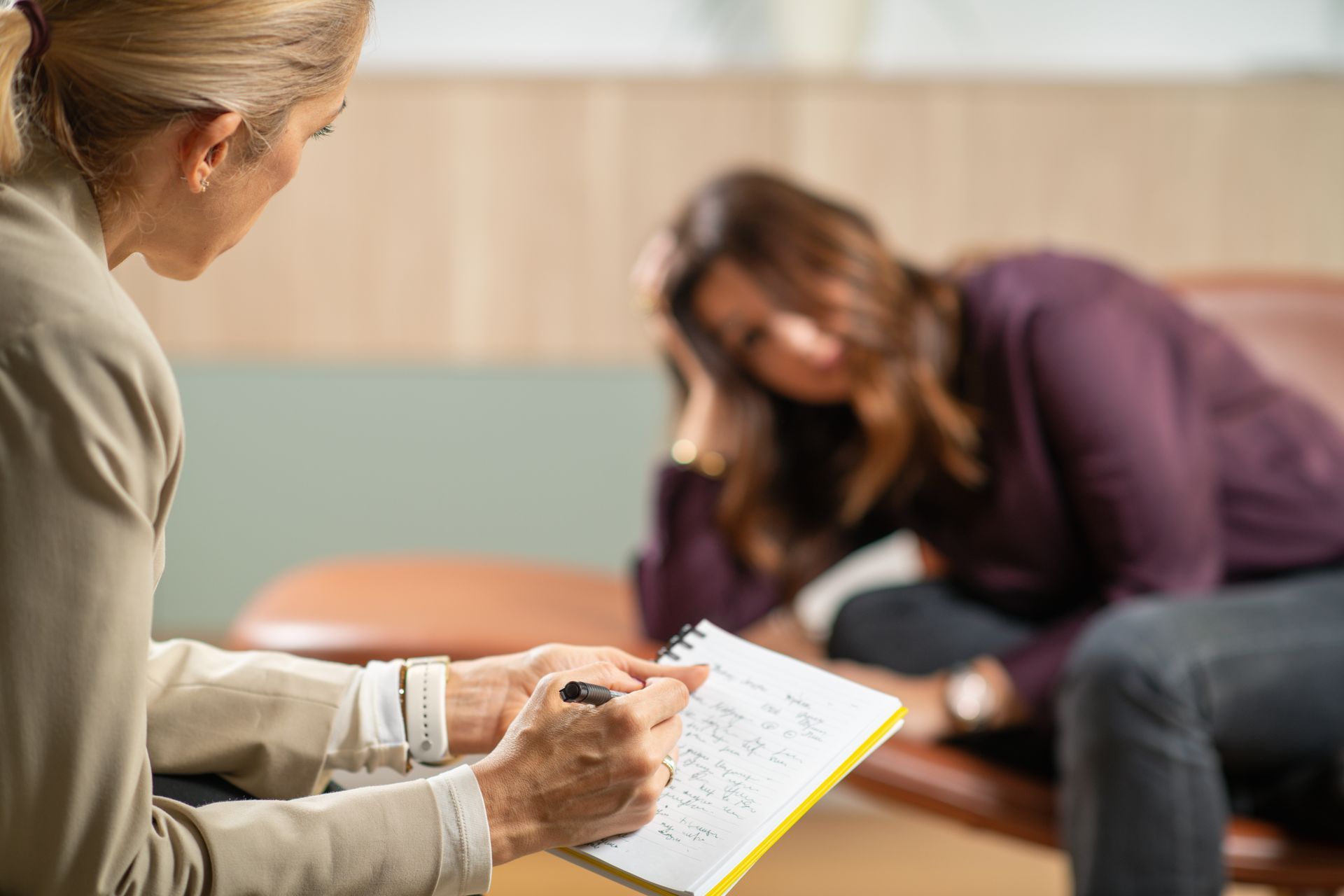 Therapist writing notes as patient looks down, resting hand on face in a consultation room.