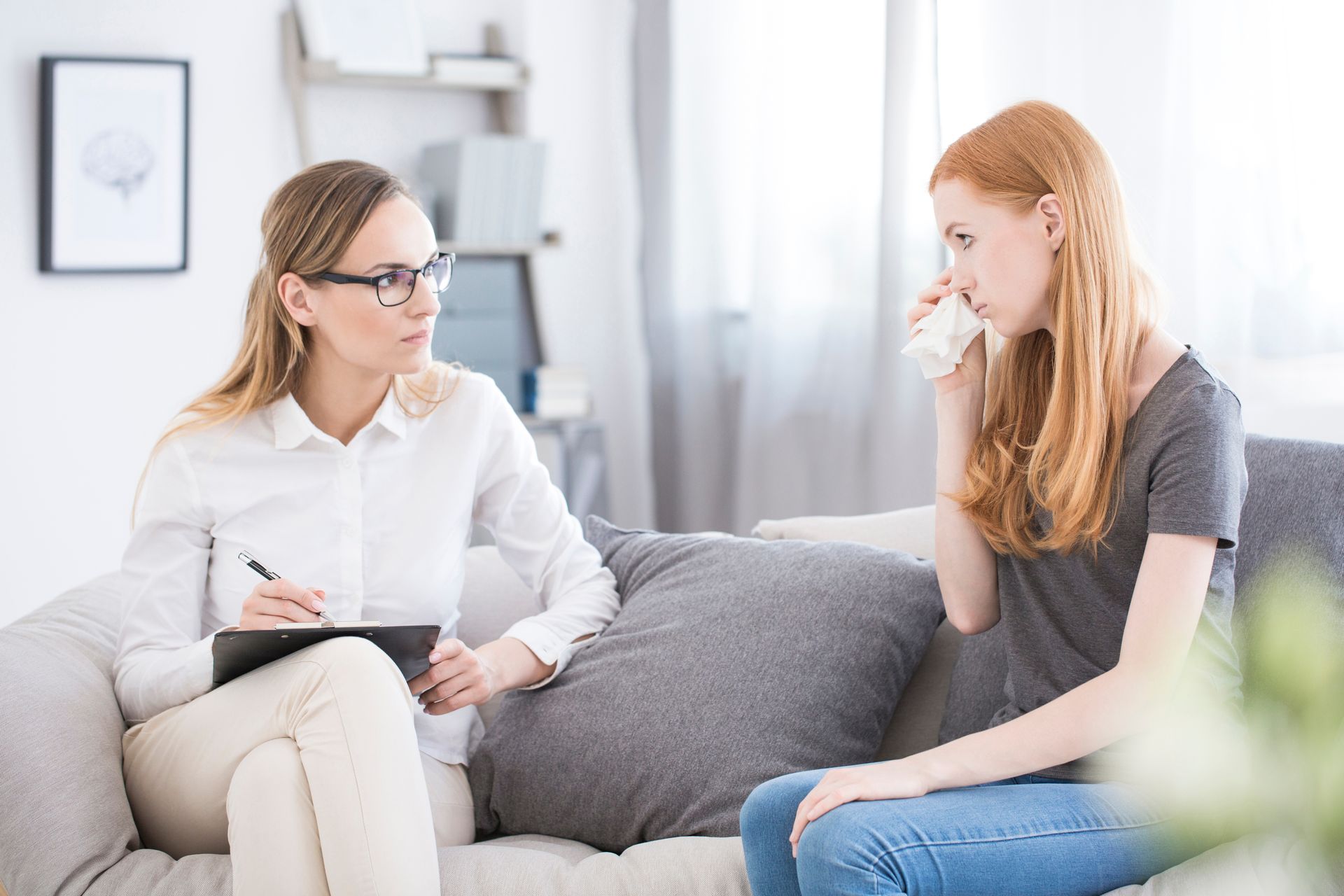 Woman crying during therapy session, therapist taking notes on couch.