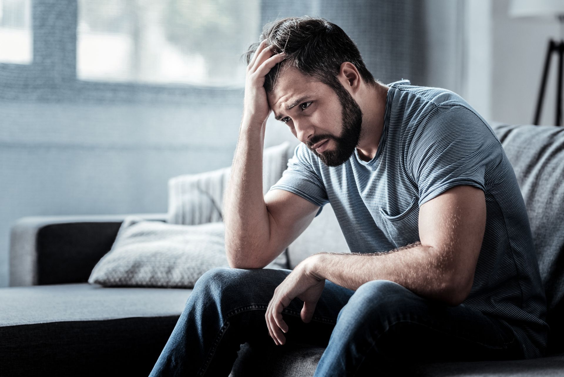 Man with hand on head, looking down with a sad expression, seated on a couch indoors.