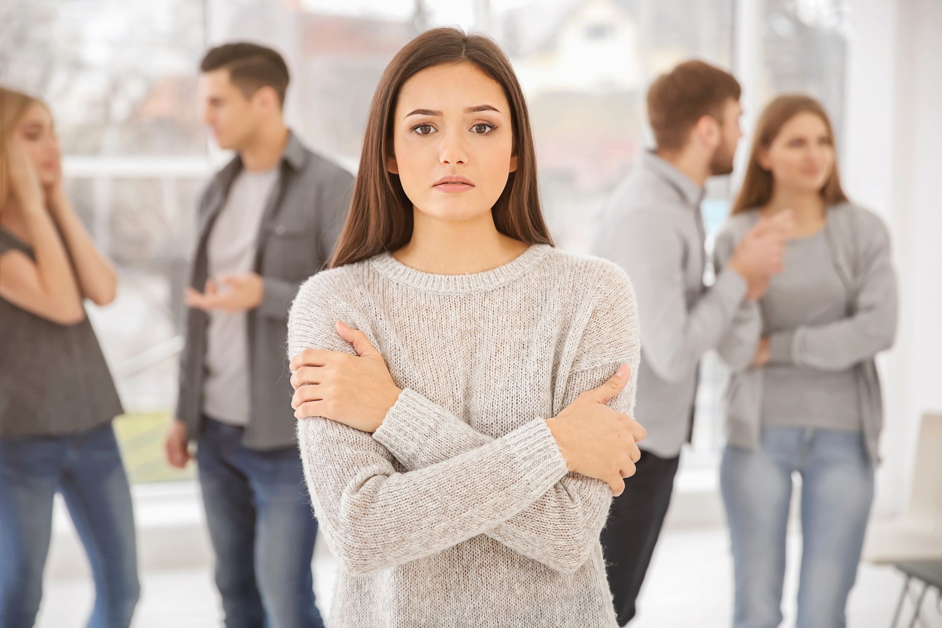Woman in a sweater looks concerned, arms crossed. People talking and looking in the background. Indoor setting.