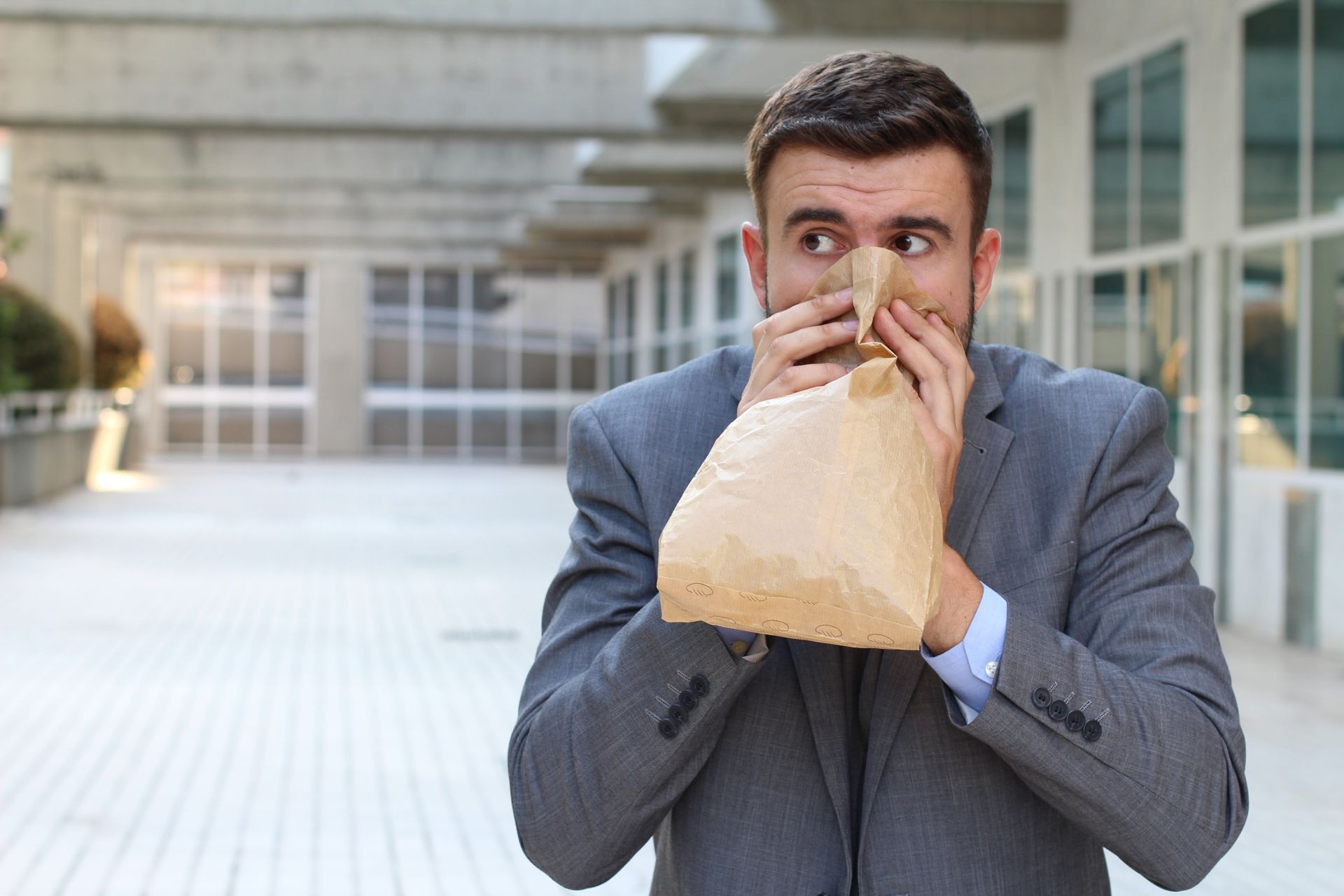 Man in suit hyperventilating into a paper bag outside a building.