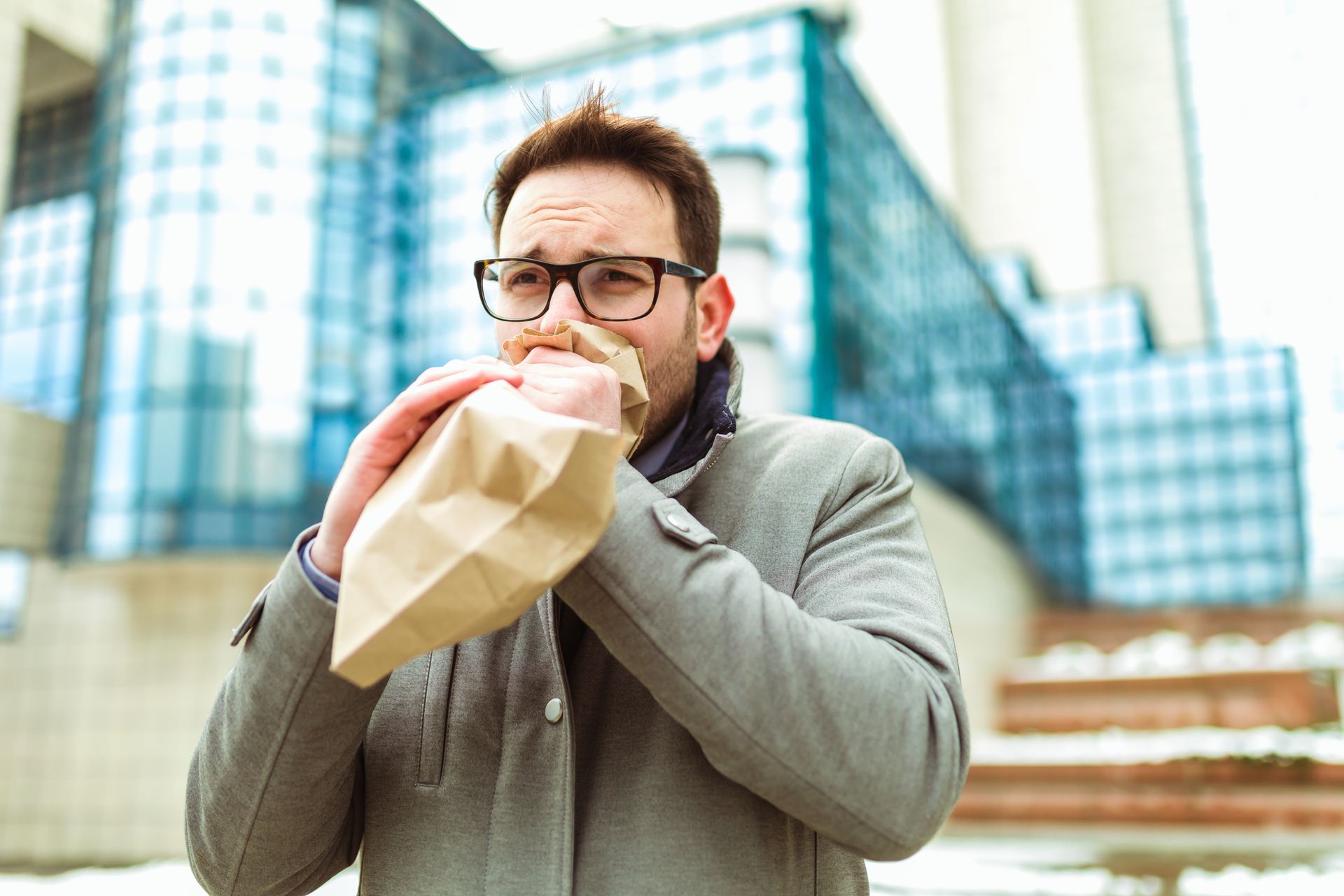 Man outside, breathing into a paper bag, likely experiencing anxiety. Glass building in background. Man outside, breathing into a paper bag, likely experiencing anxiety. Glass building in background.