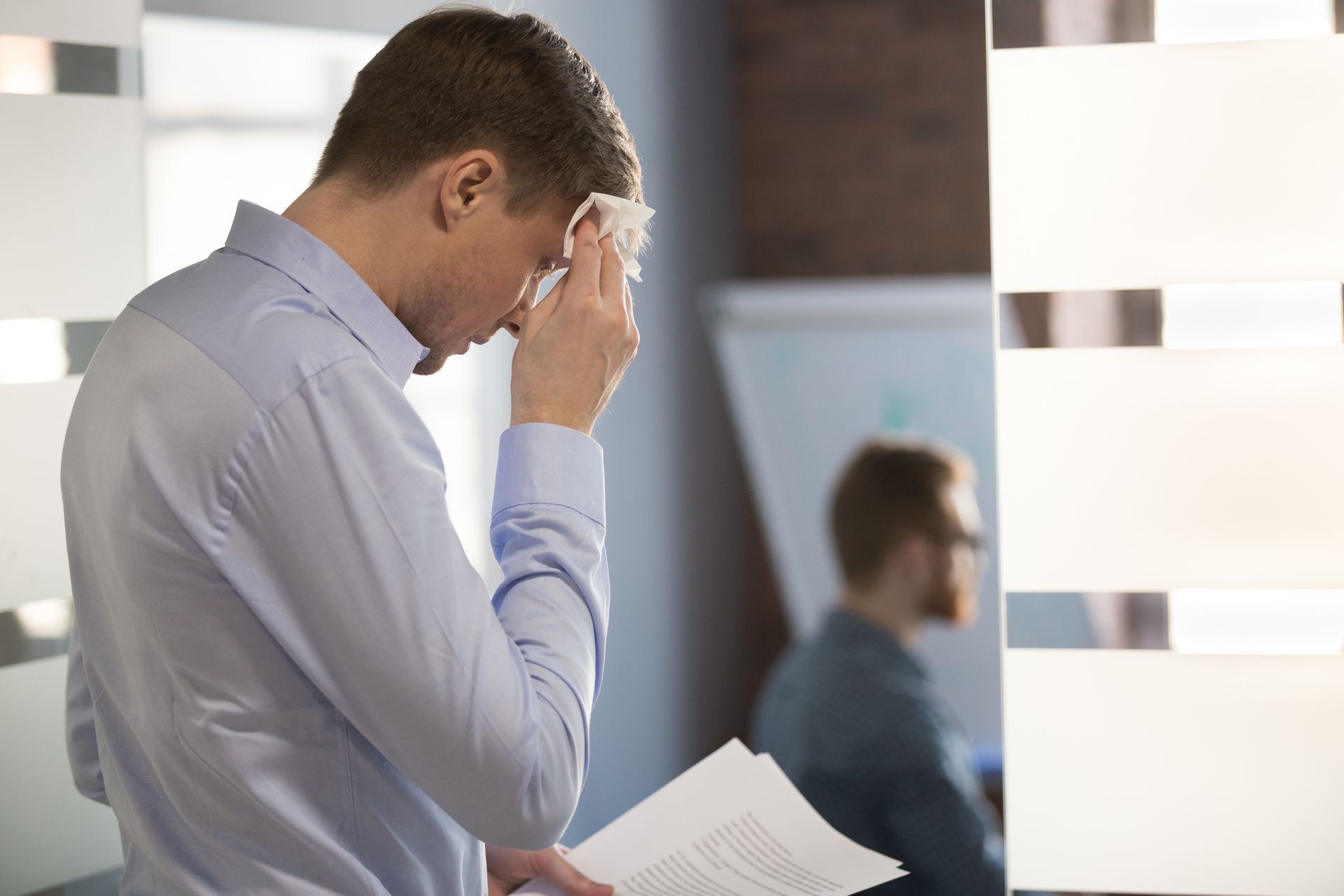Man in light blue shirt wiping sweat from forehead, holding papers; office setting.