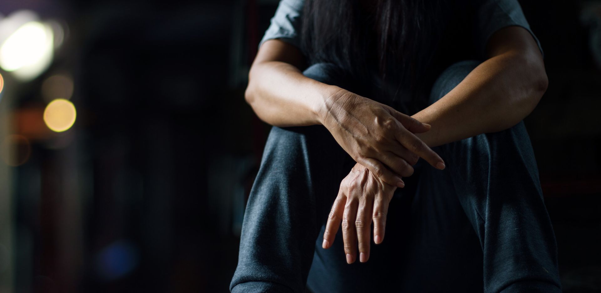 A person sitting with arms crossed, hands in focus. Dark background with blurred lights.