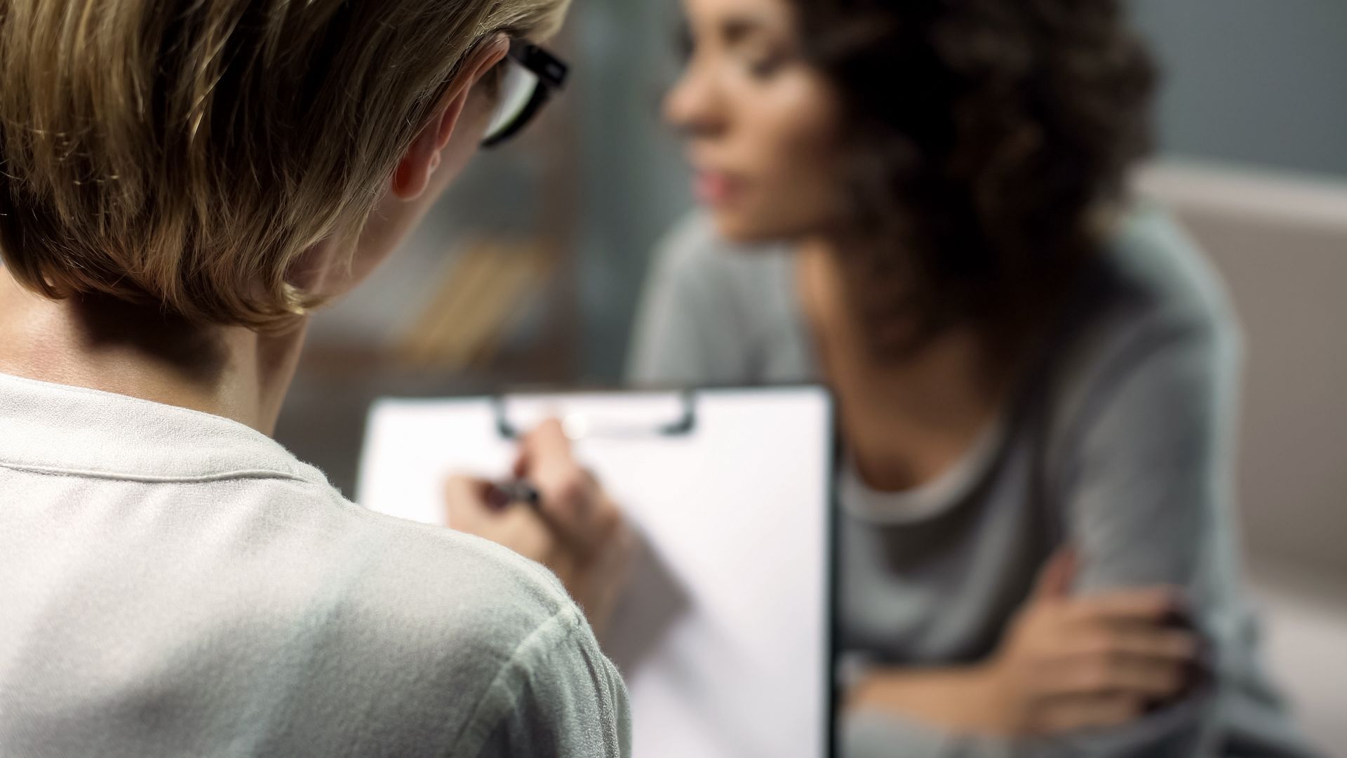 A person with glasses writes on a clipboard as another person speaks. Blurred setting suggests an office. A person with glasses writes on a clipboard as another person speaks. Blurred setting suggests an office.