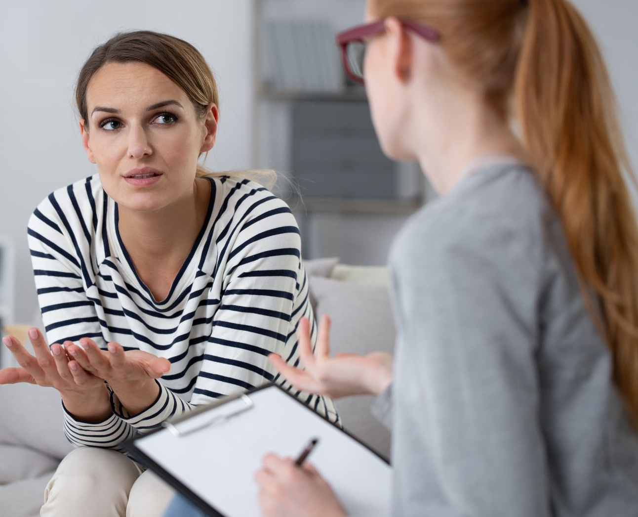 Woman in striped shirt gestures while talking to someone holding a clipboard. Indoor setting.