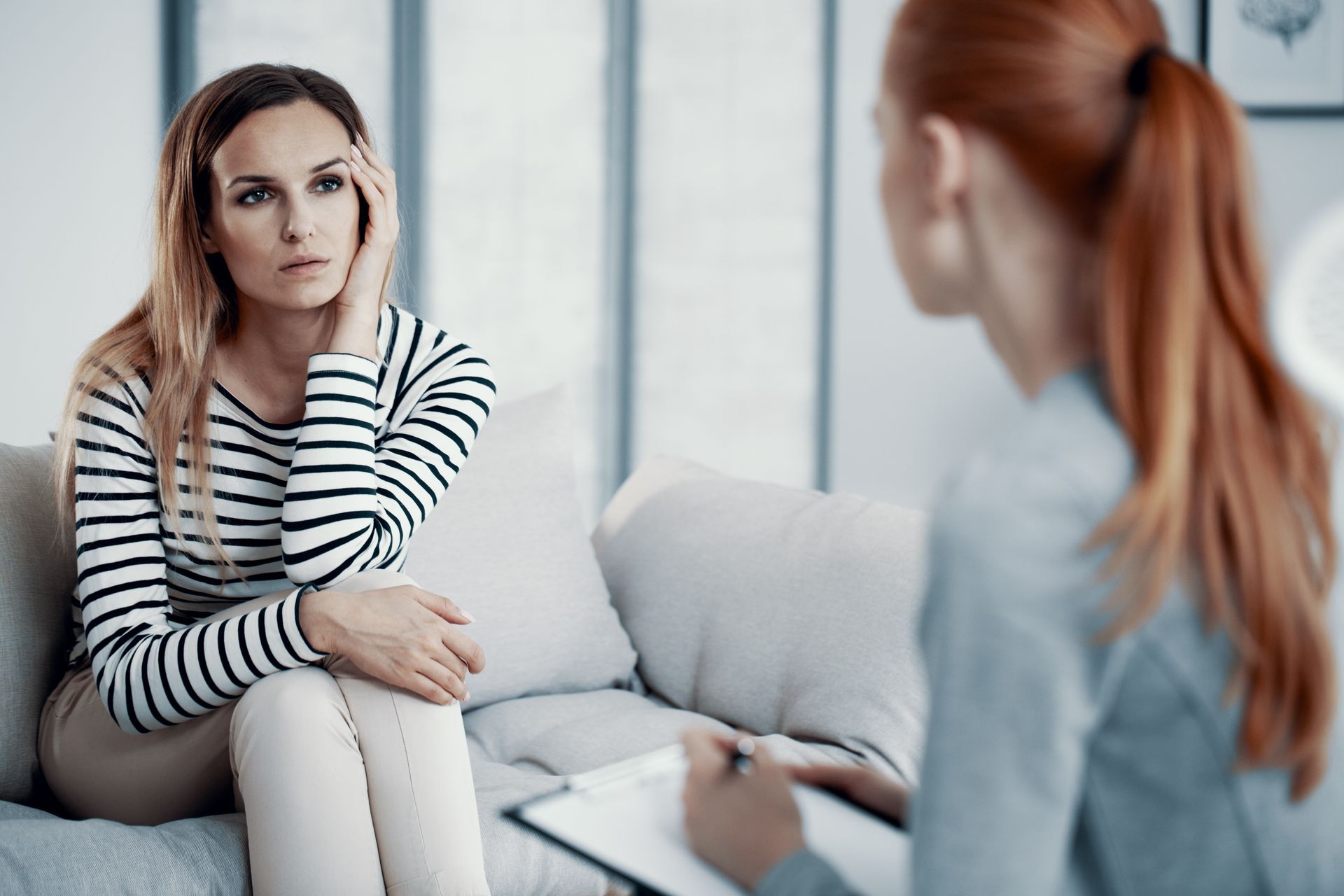 Woman on a couch with concerned expression, talking to a therapist holding a clipboard in a well-lit office.
