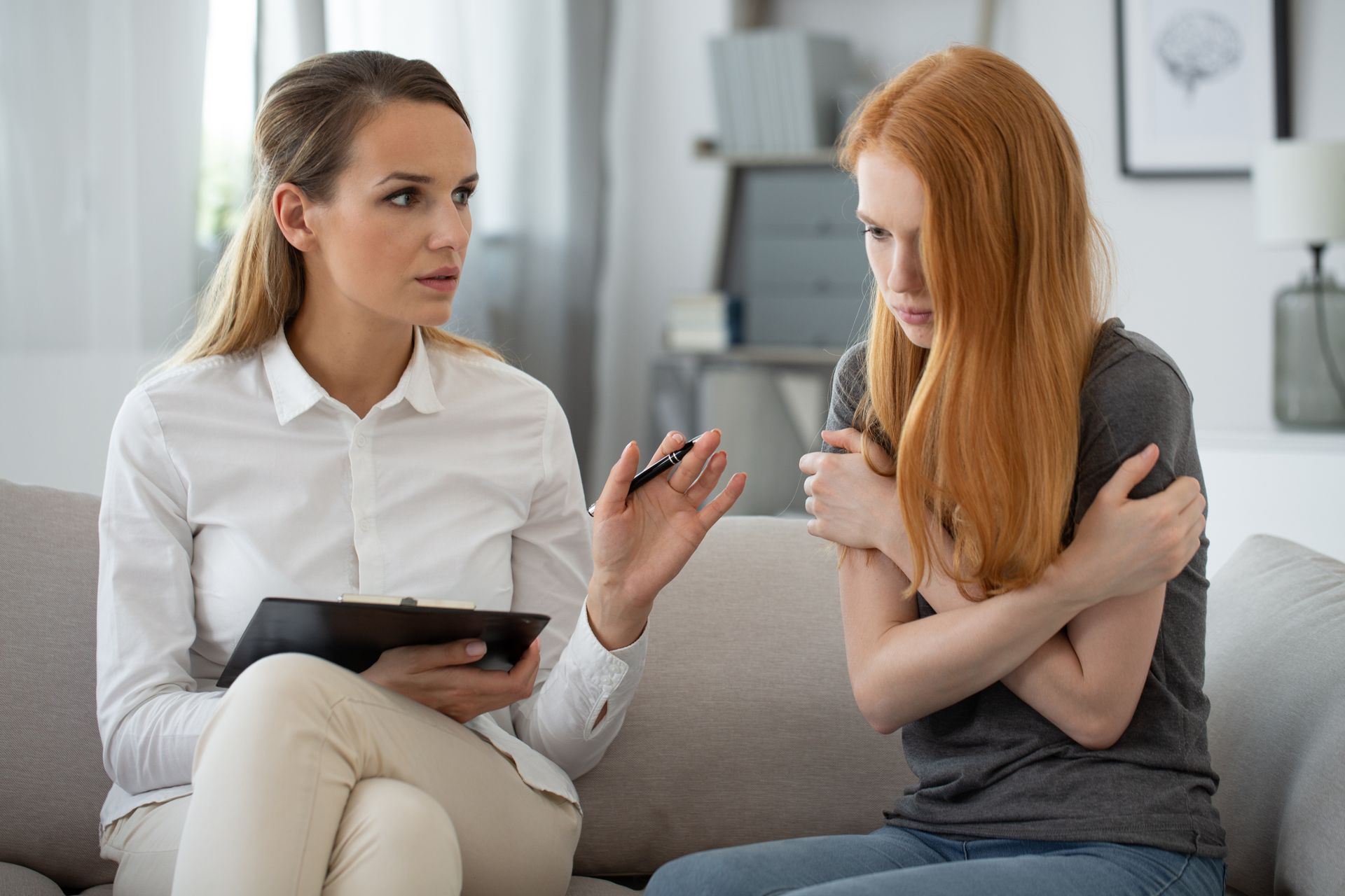 Woman in white shirt with notepad talking to a woman, arms crossed, looking concerned. Living room.
