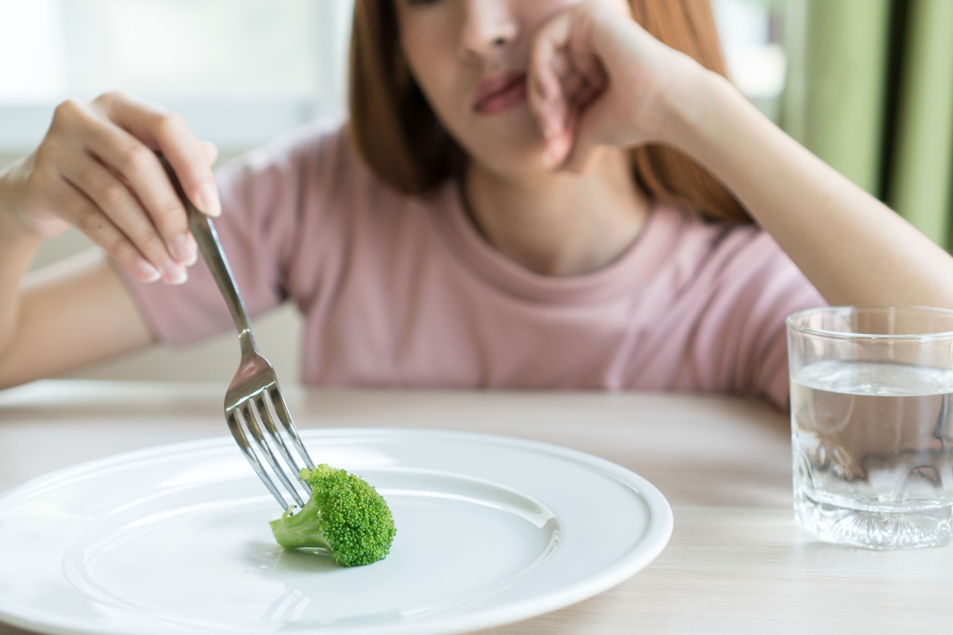 Woman with fork, broccoli on plate, looking weary, next to glass of water.