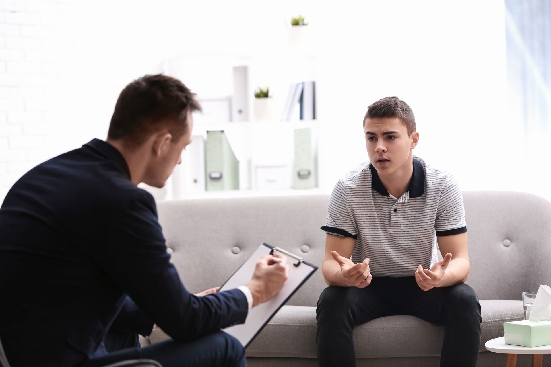 Man in suit taking notes while teenager talks on sofa in a brightly lit counseling session room.
