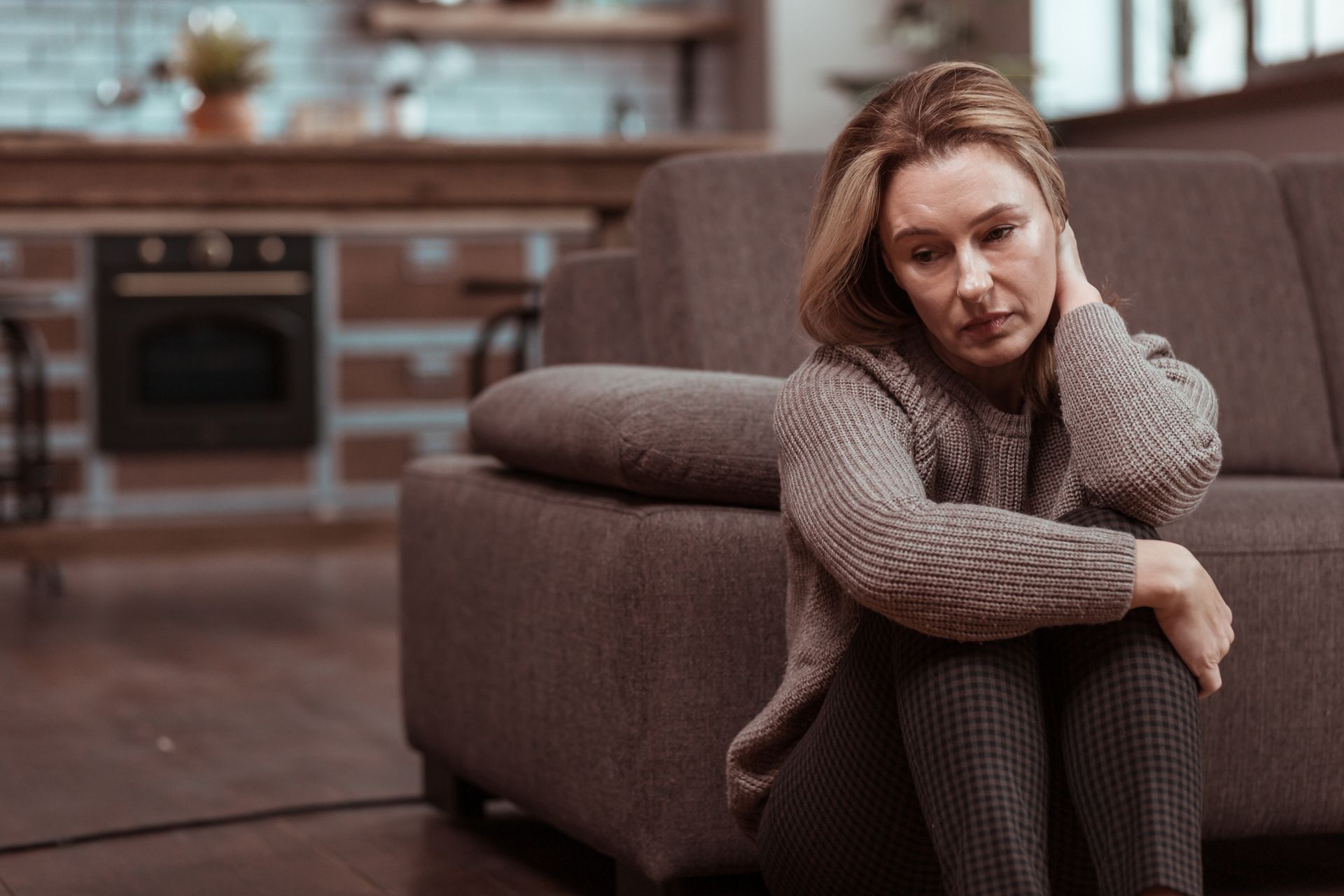 Woman sitting on floor next to sofa, looking down with a sad expression. Cozy home setting.